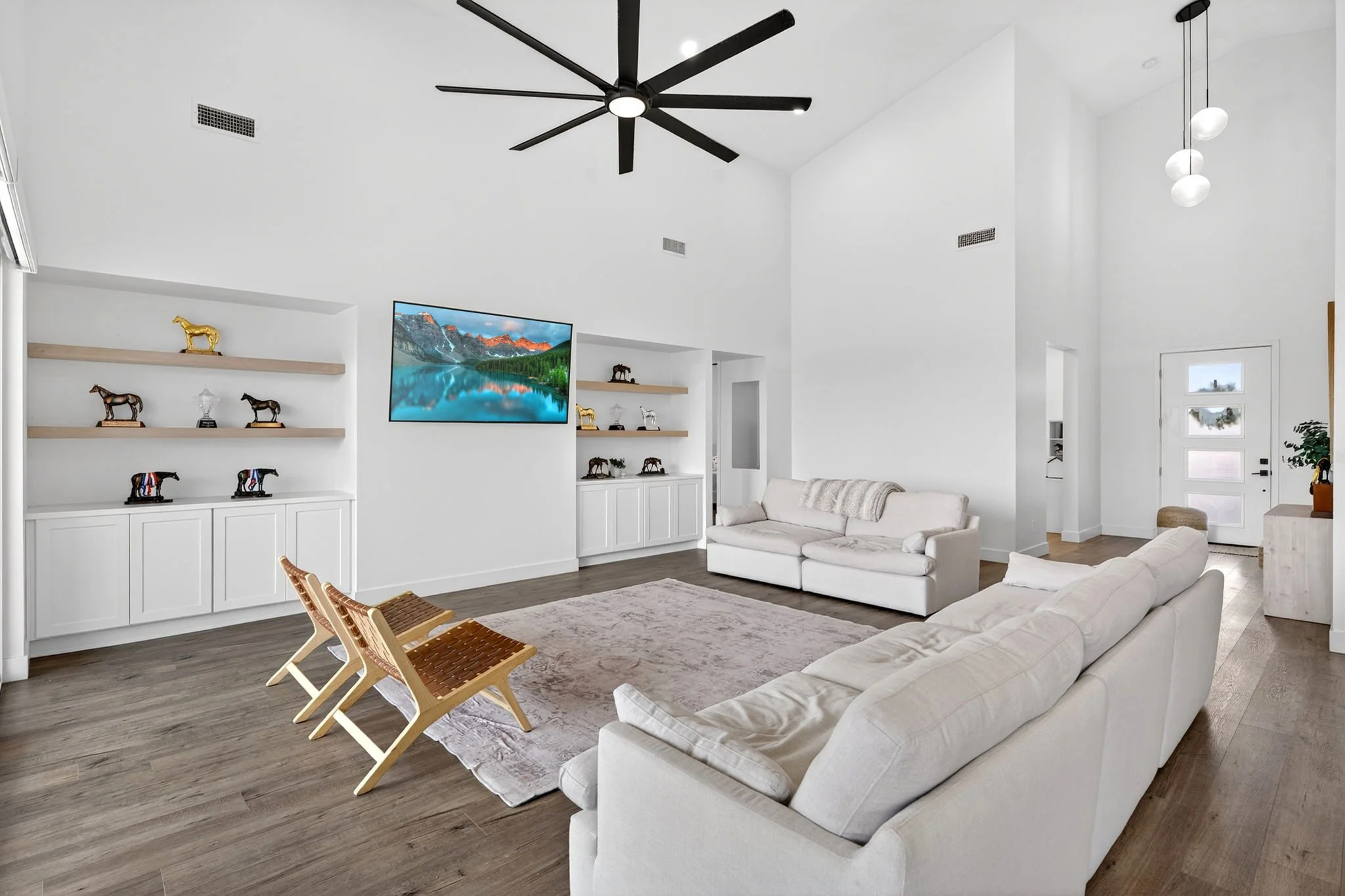 Living room with white walls and ceiling, black ceiling fan, modern pendant lights, white sofas, wooden chairs, built-in shelves with decorative horse figurines, mounted flat-screen TV showing mountain scenery, hardwood floor, and door with frosted g