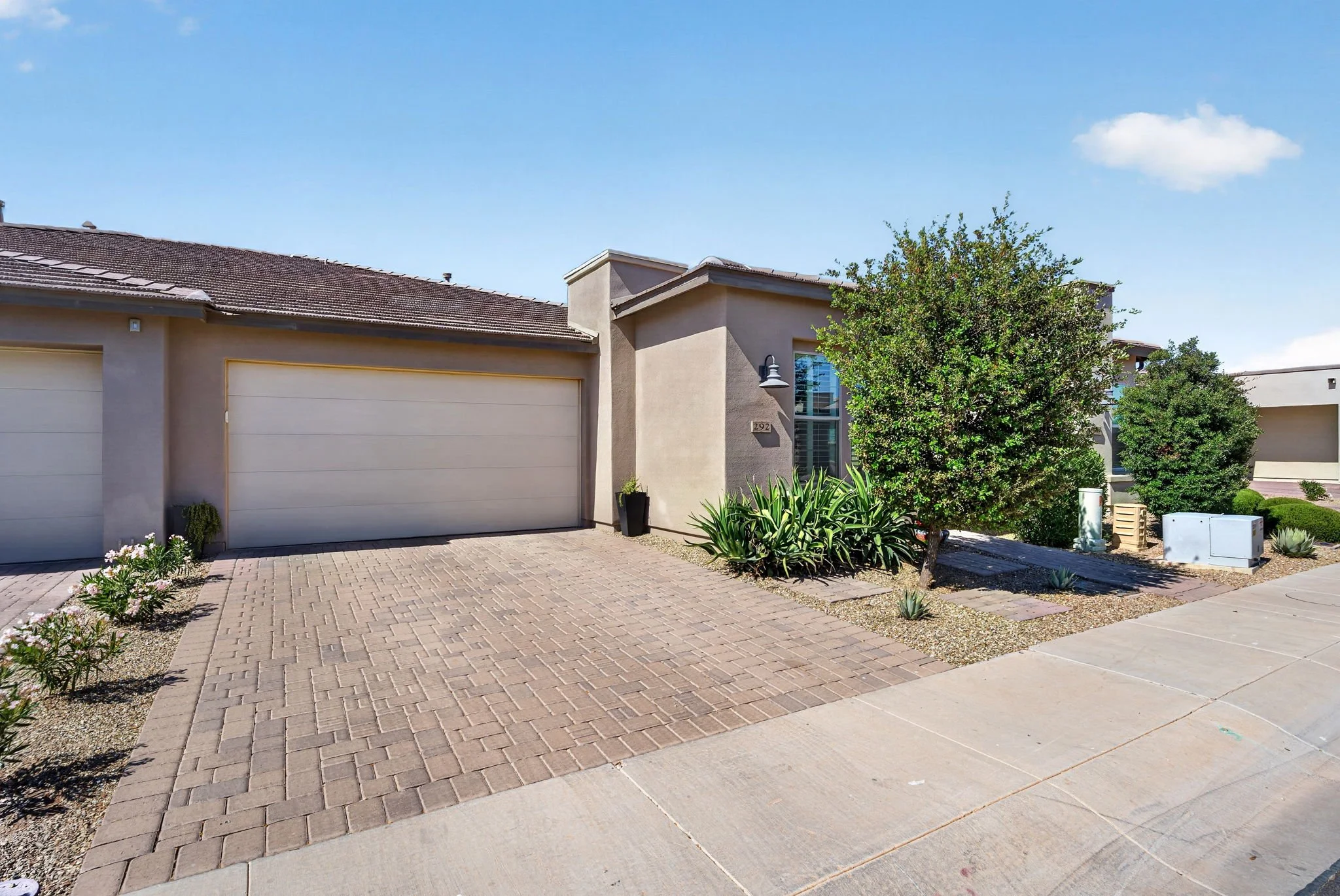 A modern house with a beige exterior, a two-car garage with white doors, a brick-paved driveway, small shrubs, and a large tree in front, under a clear blue sky.