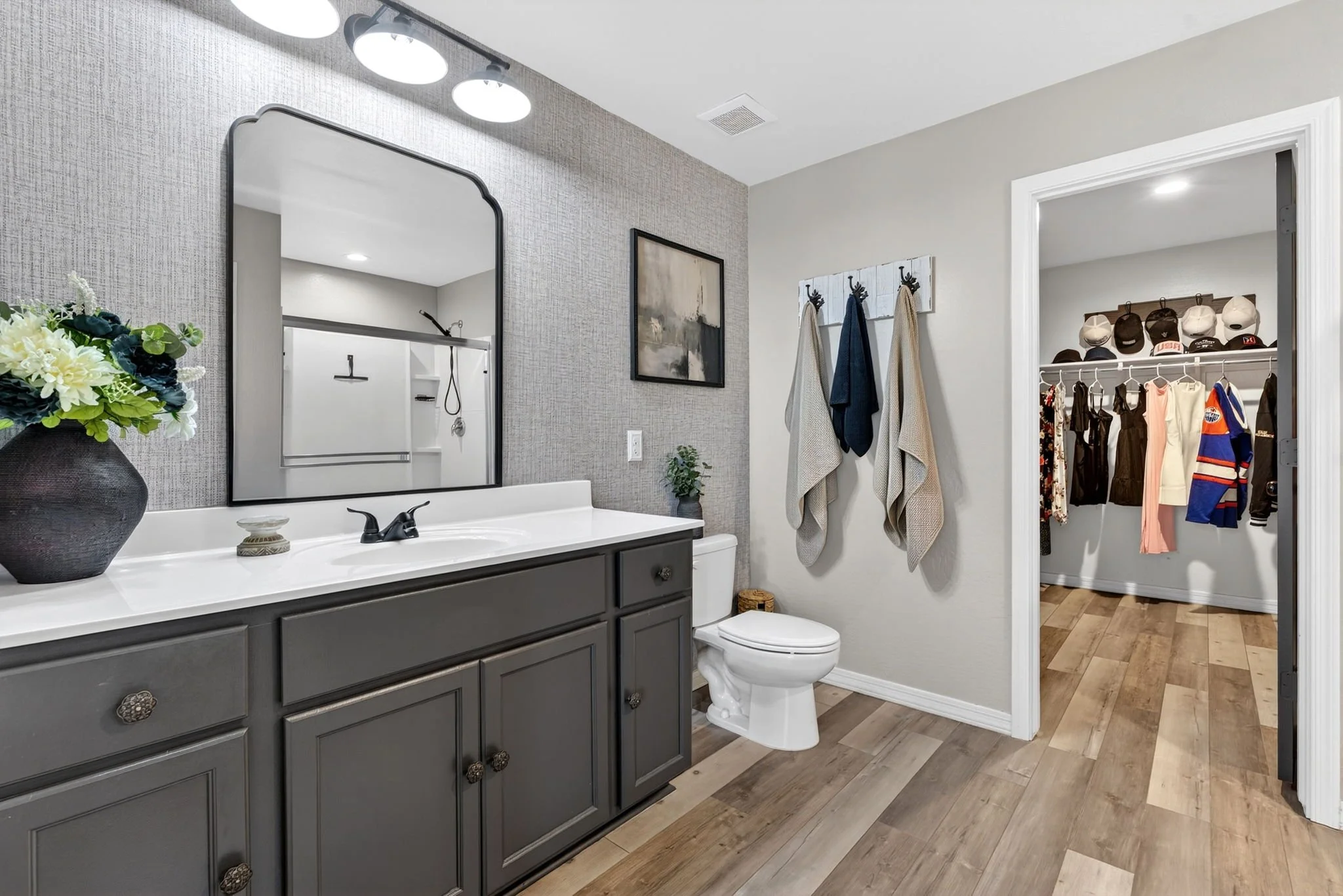 A modern bathroom with a gray vanity with decorative knobs, a large mirror, a white countertop, and a black faucet. There is a vase with flowers on the counter. Adjacent to the bathroom is a walk-in closet with hanging clothes, hats, and hooks on the