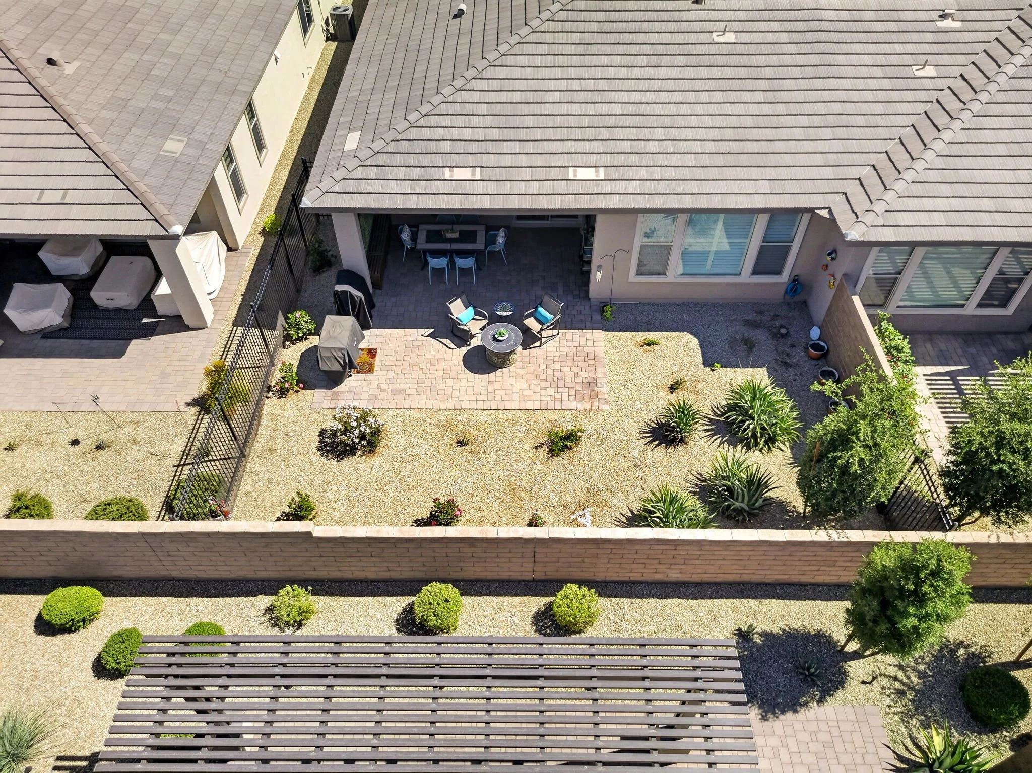An aerial view of a backyard patio area with outdoor furniture, including chairs and a table, surrounded by a fenced yard with plants and shrubs.