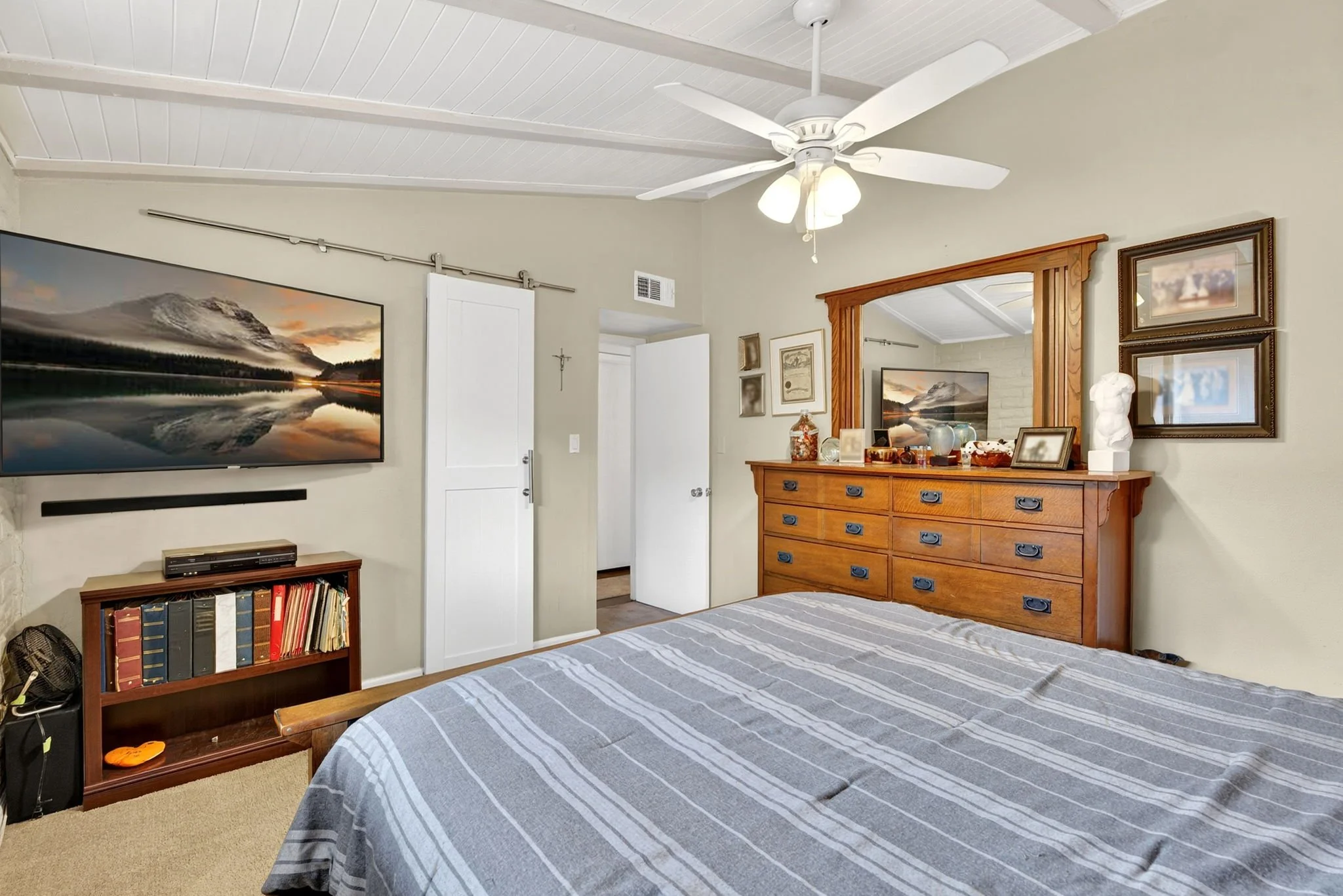 A cozy bedroom with a bed covered in a gray-striped blanket, a wooden dresser with decorative items, and wall art. An open door reveals another room, and a flat-screen TV is mounted on the wall.
