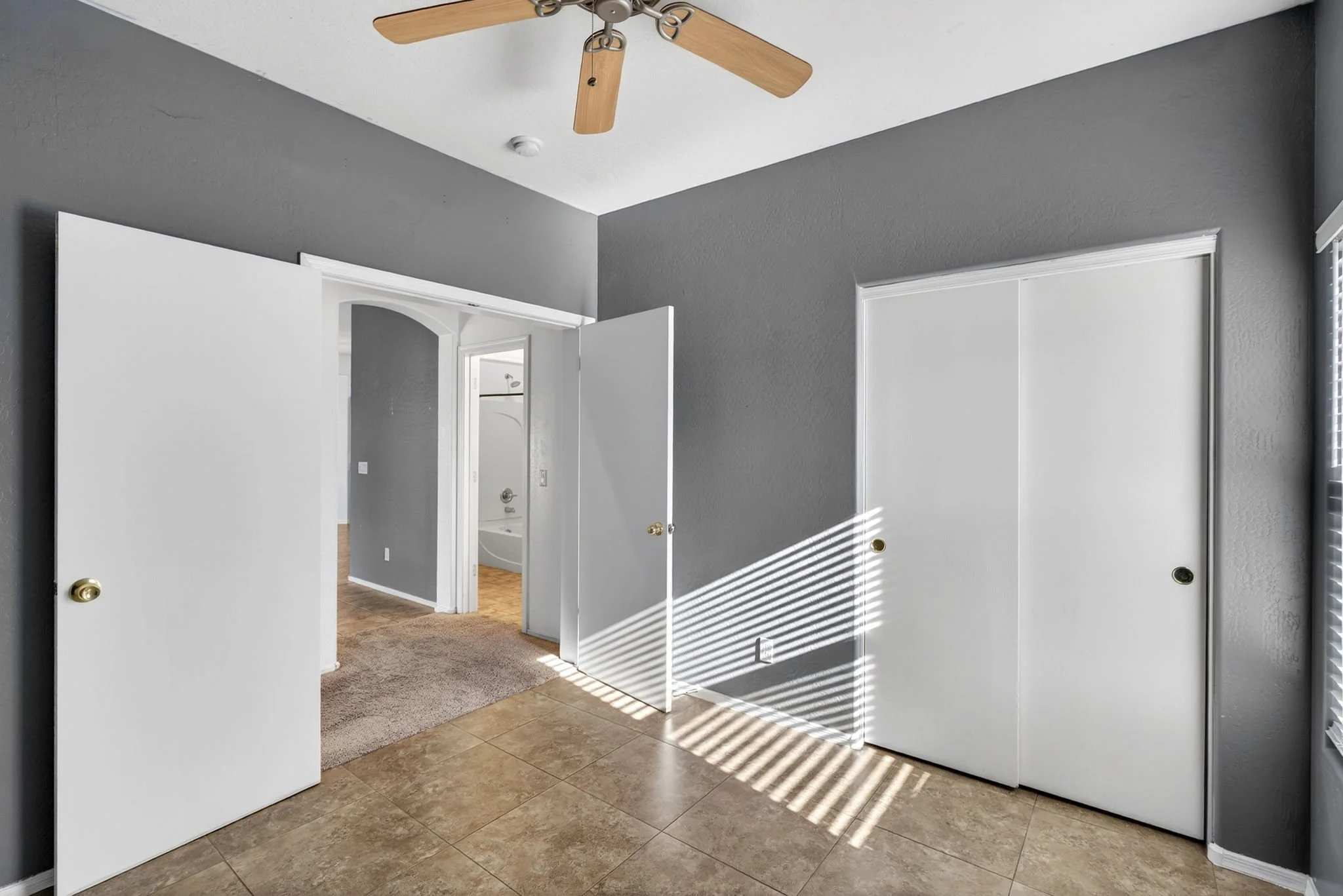 Interior view of a room with open white doors, gray walls, a ceiling fan with wooden blades, tiled and carpeted flooring, and sunlight creating shadows on the wall.