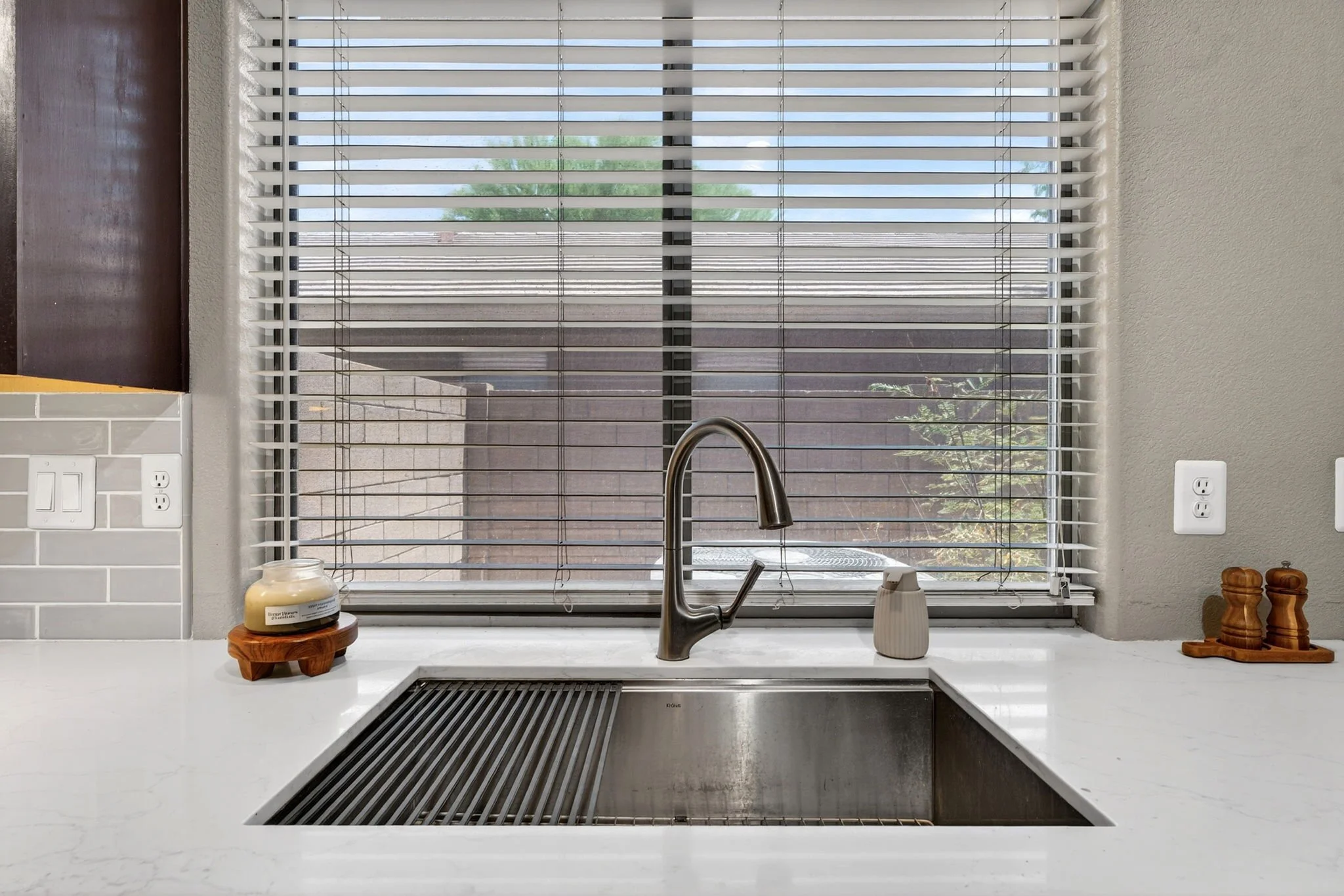 A kitchen window with white horizontal blinds, view of trees outside, granite countertop in the foreground, a stainless steel faucet, and a soap dispenser.