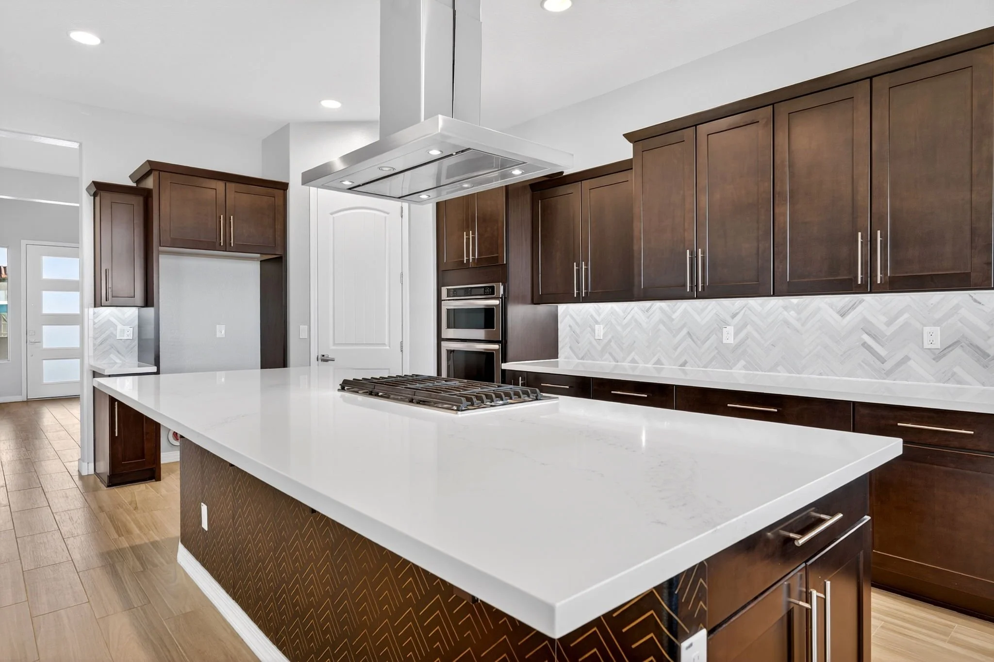 Modern kitchen with dark wood cabinets, white countertops, and a stainless steel range hood above a stove, with a white chevron backsplash and hardwood flooring.