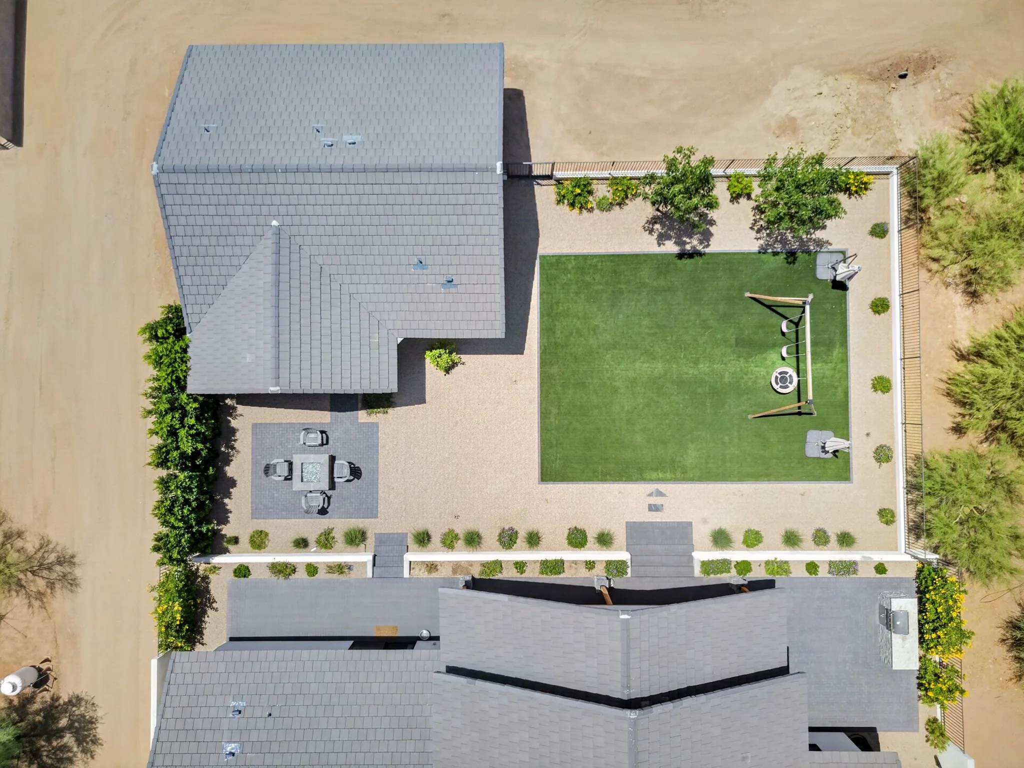 An aerial view of a residential backyard featuring a gray-roofed house, a green artificial turf area with a swing set, and mature trees along the fence. Adjacent to the house is a gravel area with outdoor furniture and a surrounding white fence with 