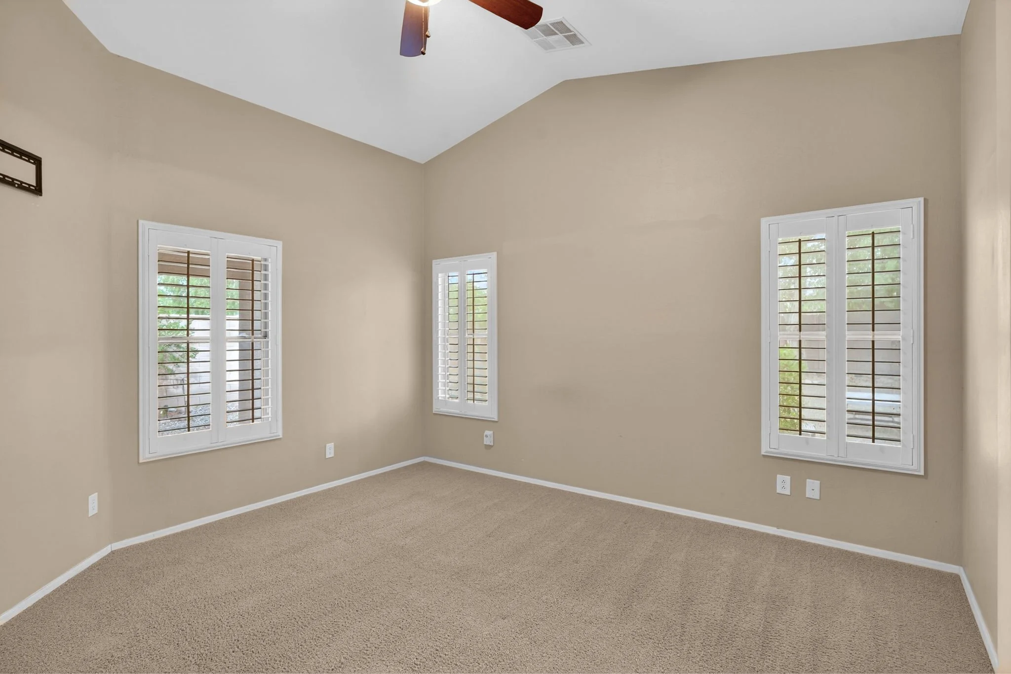 Empty room with beige walls, beige carpet, four windows with white shutters, and a ceiling fan.