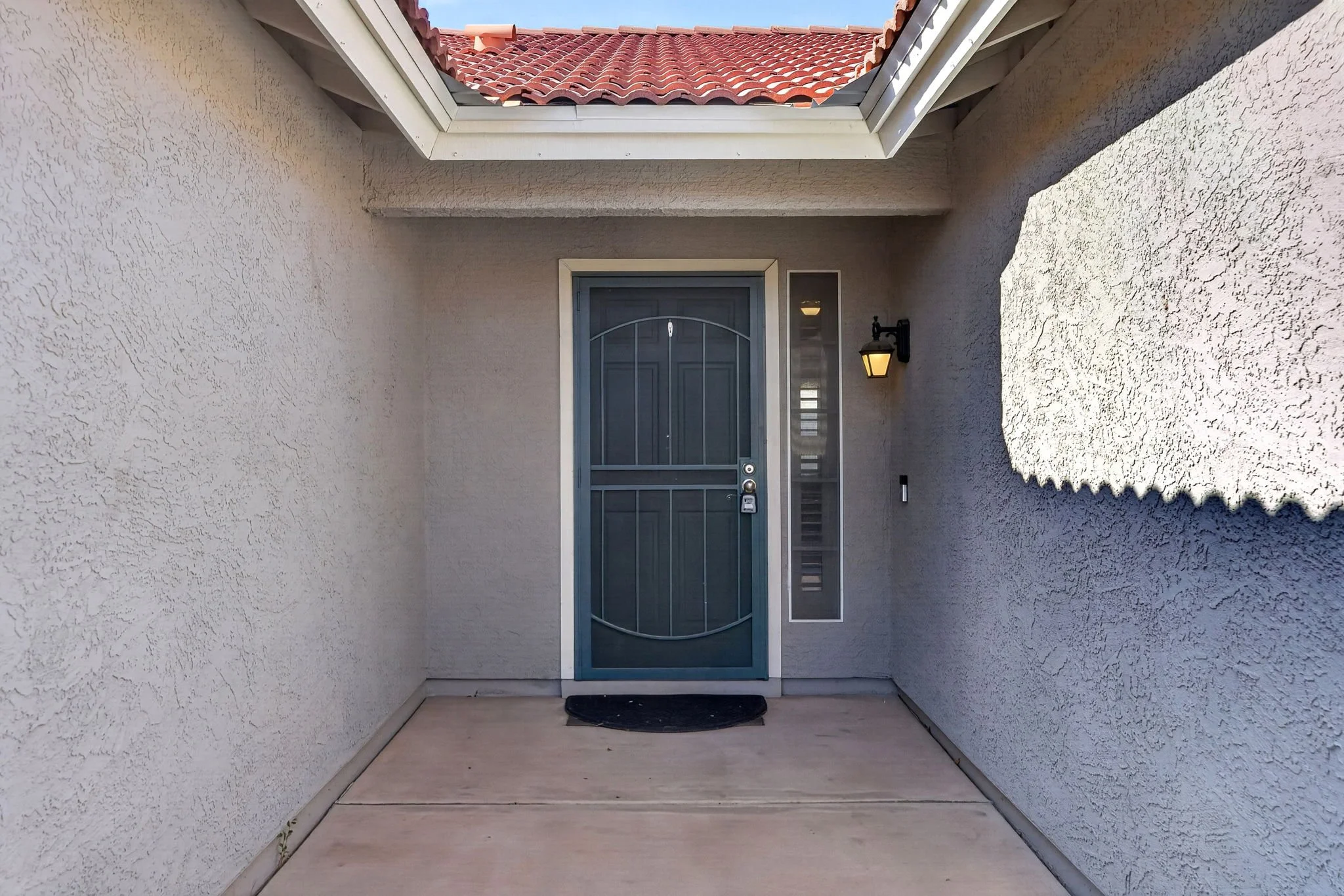 Front porch entryway with a blue door, stucco walls, a window to the right, a black outdoor wall lantern, and a red tile roof visible above.