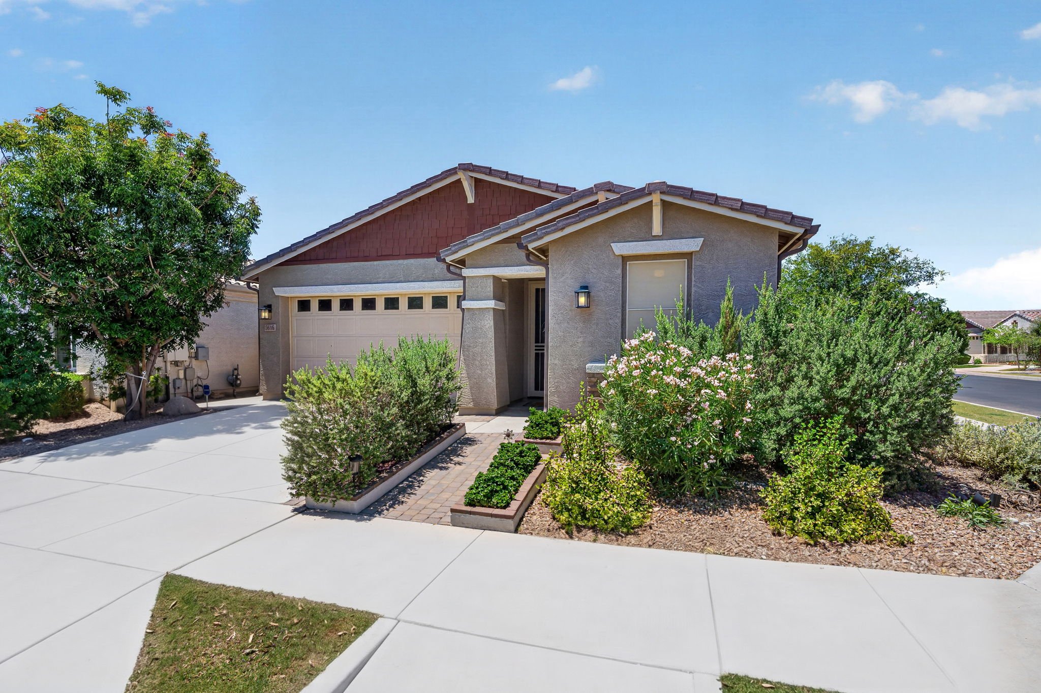 A suburban house with a two-car garage, beige exterior, and a front yard with bushes and a small garden. There are trees and a blue sky with some clouds in the background.