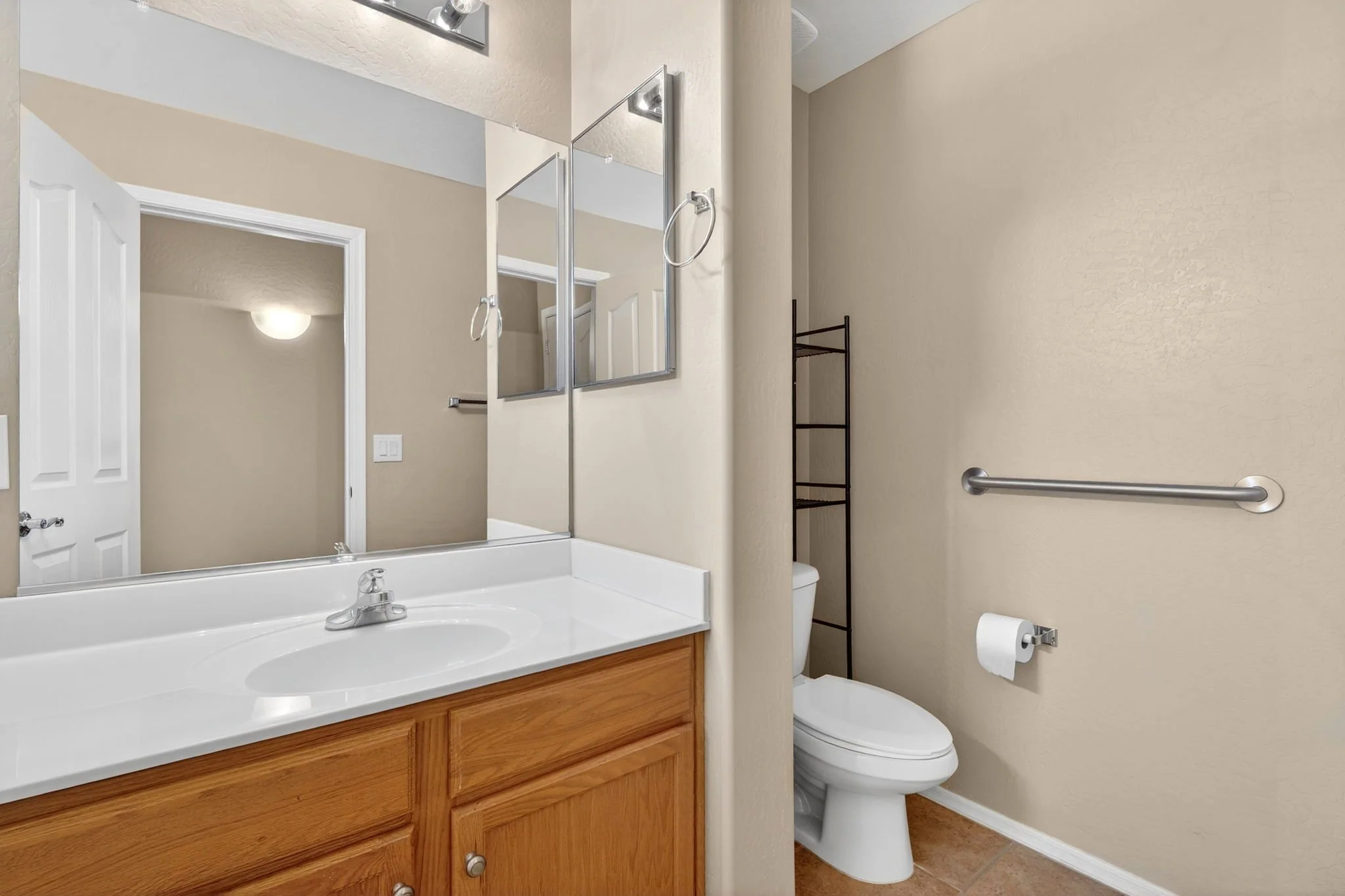 A small bathroom with a vanity sink, wooden cabinet, and a mirror. There is a toilet with a grab bar on the wall beside it, a black metal shelving unit, and a beige painted wall.