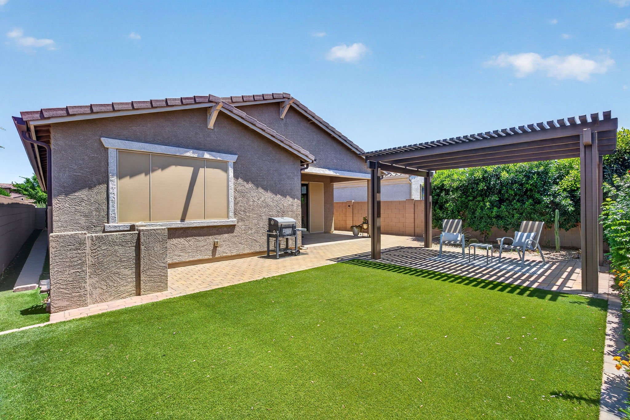 A backyard patio with a paver floor, an artificial grass patch, a covered seating area with chairs, a grill, and a house wall with a window, under a blue sky with a few clouds.