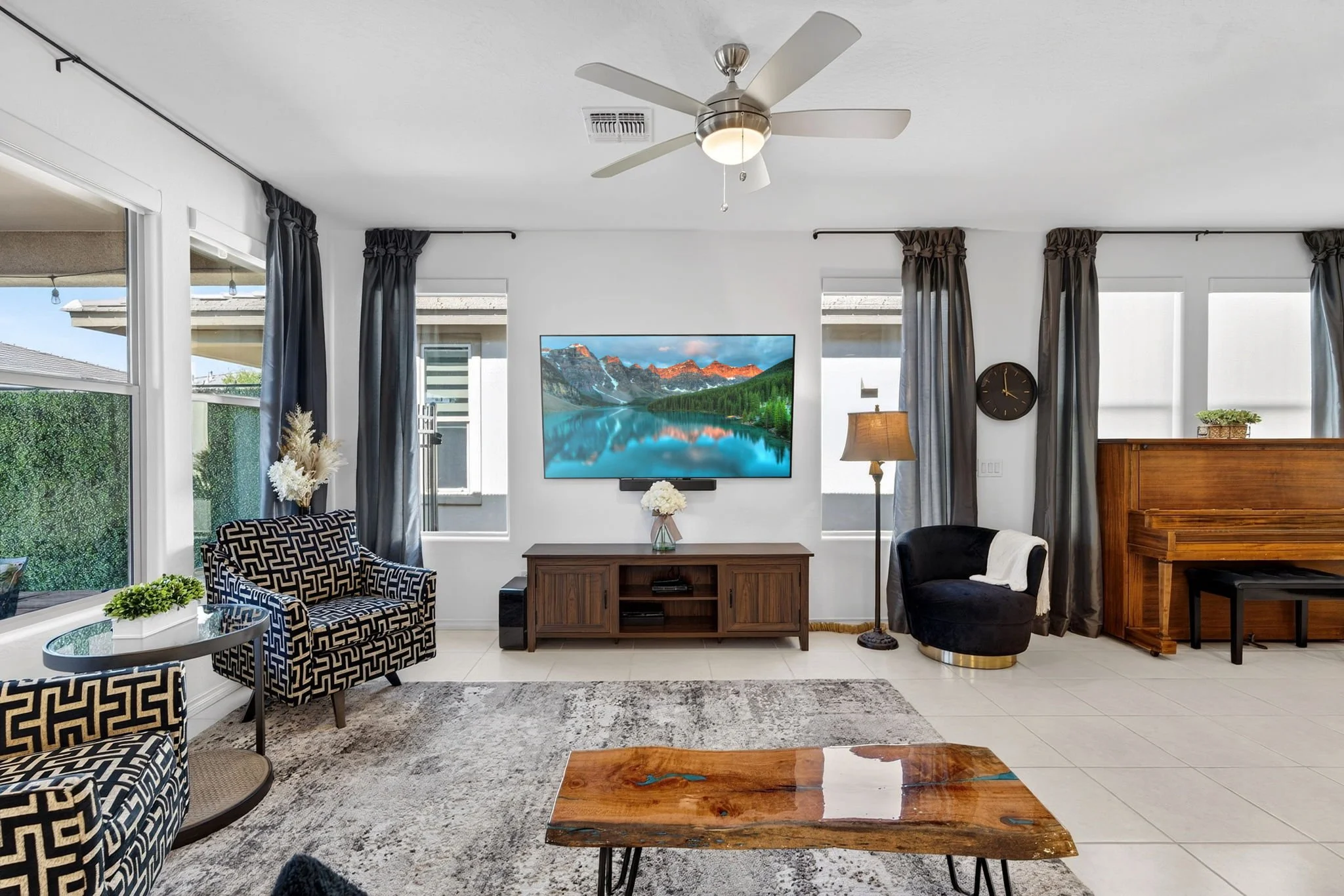 Living room with large windows, black and white patterned chairs, wooden coffee table, TV mounted on wall, ceiling fan, floor lamp, and piano.