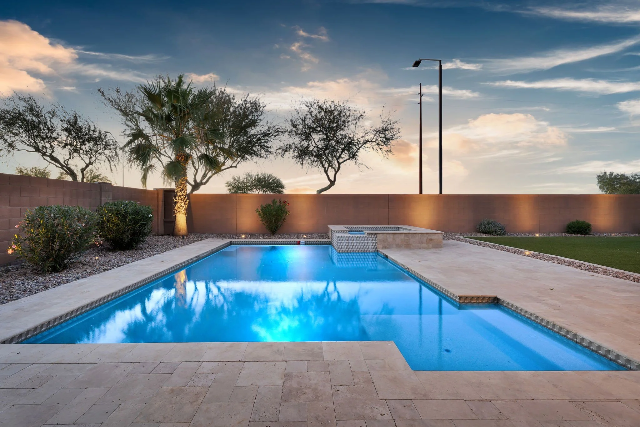 Backyard with a rectangular swimming pool, surrounded by stone pavers, landscaping, and a brick wall, with trees and a sky with clouds at sunset.