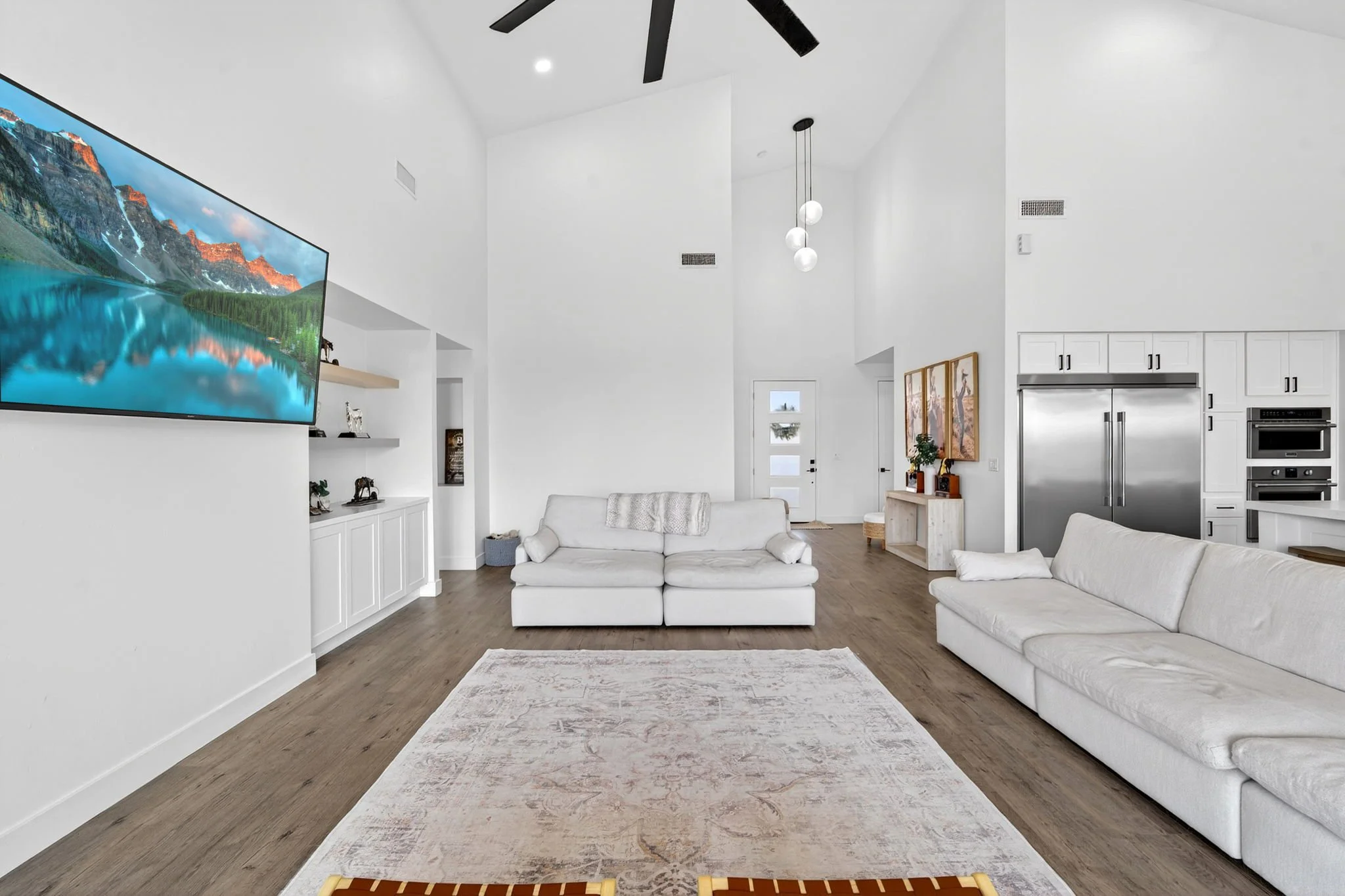 Living room with white sofas, a large wall-mounted TV displaying a mountain landscape, a light-colored rug on wood flooring, a hanging light fixture with three globes, and kitchen cabinets with stainless steel appliances.