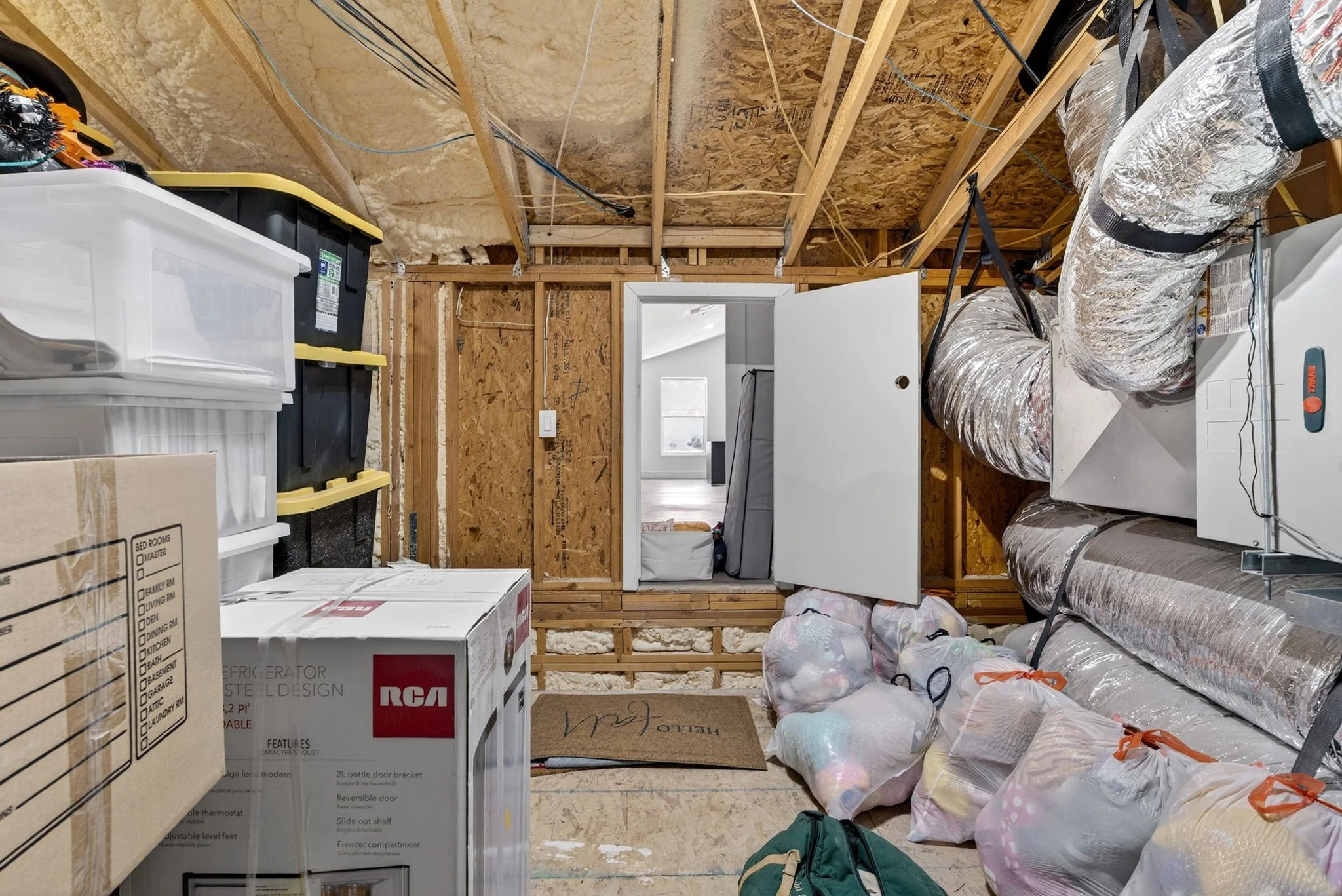 Empty unfinished room with exposed wooden framing, insulated walls, ductwork, bins, boxes, and household items packed in plastic bags.