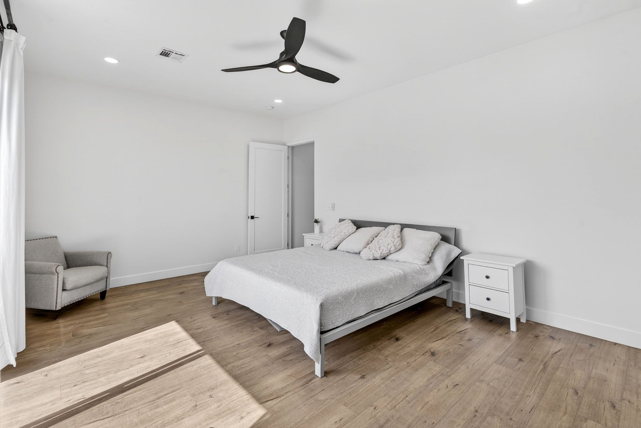A minimalist bedroom with a white bed, nightstand, and an armchair, with a ceiling fan and recessed lighting.