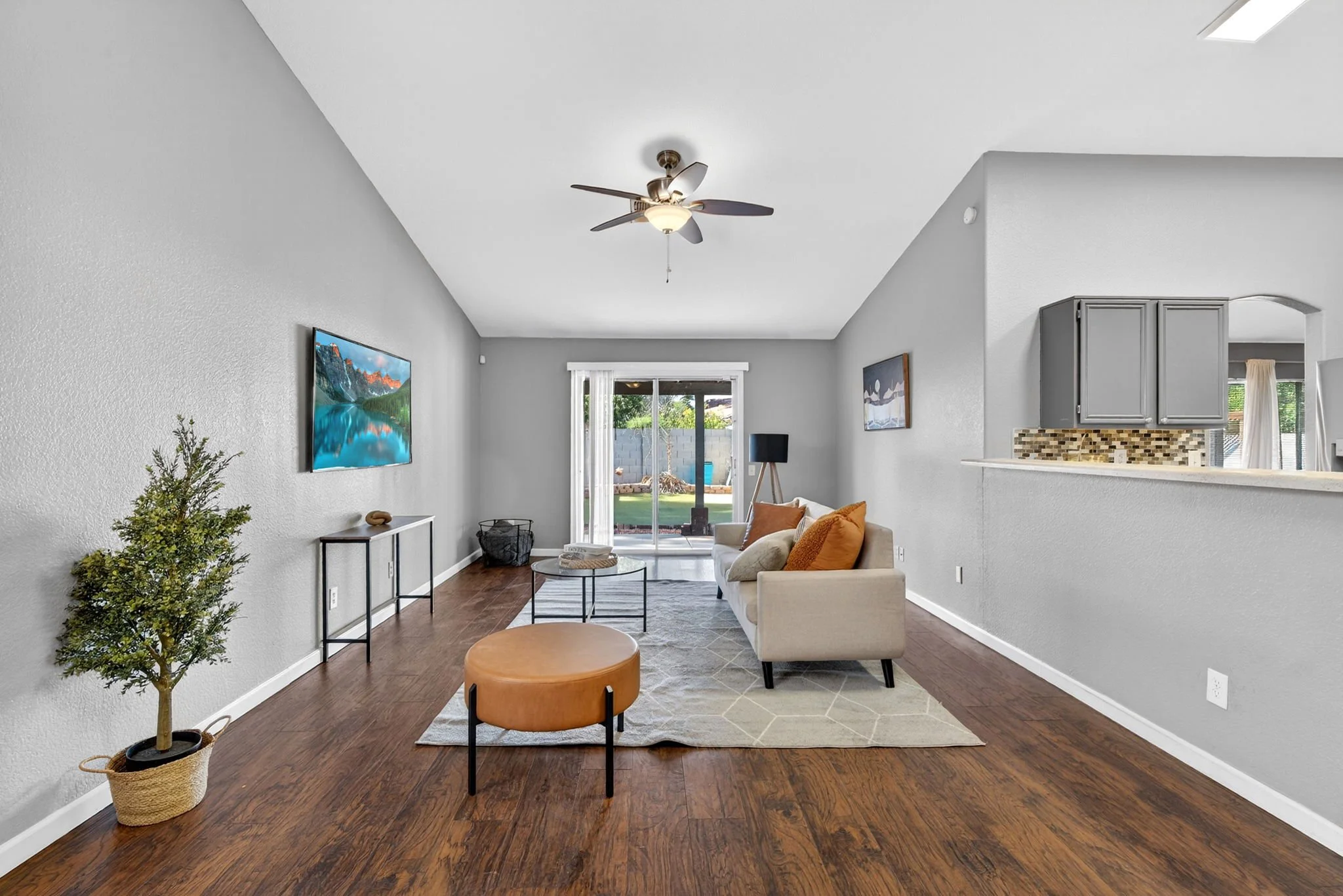 Living room with a white sofa, orange pillows, a glass coffee table, a green potted plant, a wall-mounted TV, and a sliding glass door leading to the backyard