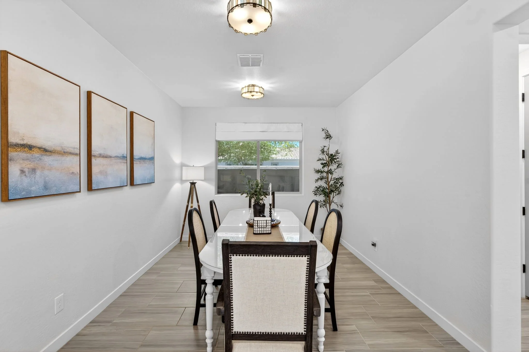 A bright dining room with white walls, a window with a white blind showing green trees outside, and three ceiling lights. There is a white dining table with six chairs, a table runner, and a centerpiece. Artwork and a floor lamp are on the walls, and