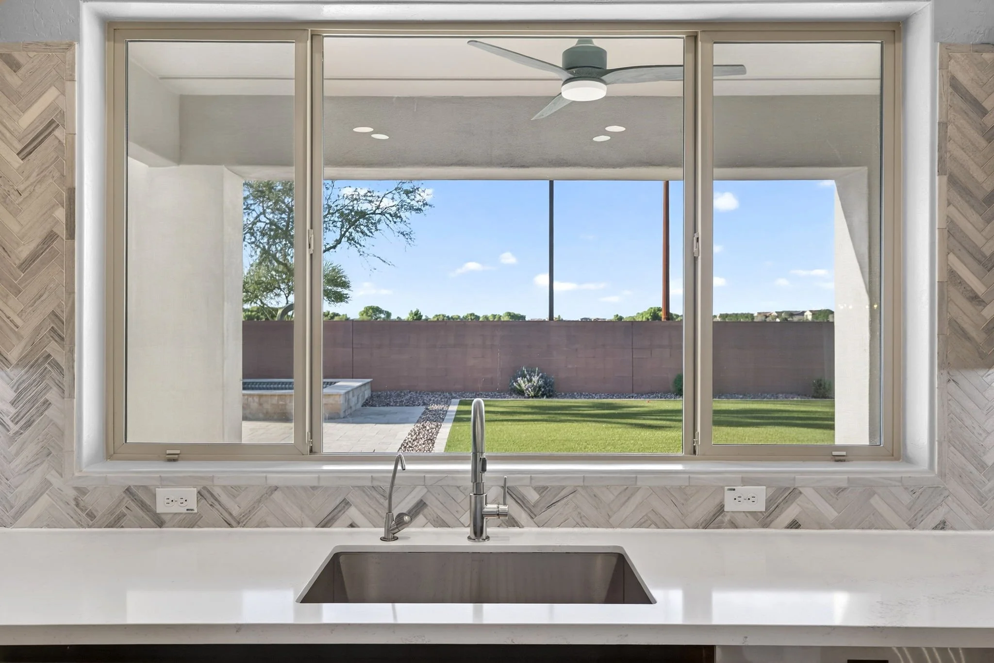 Kitchen sink with a view of a backyard through large window, ceiling fan, and blue sky.