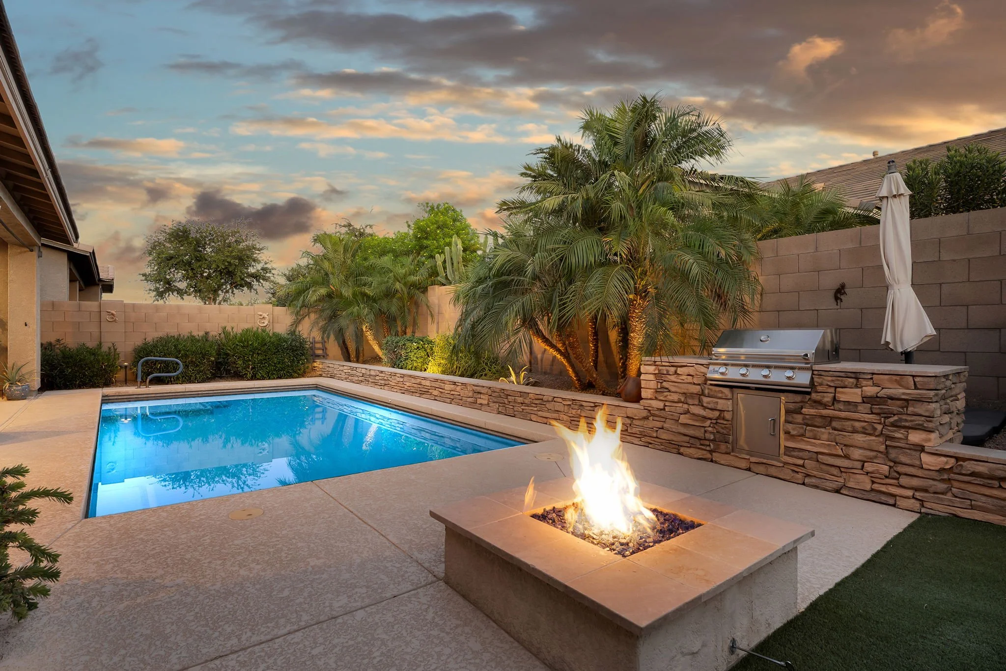 A backyard with a swimming pool, a fire pit, palm trees, and a barbecue grill at sunset.