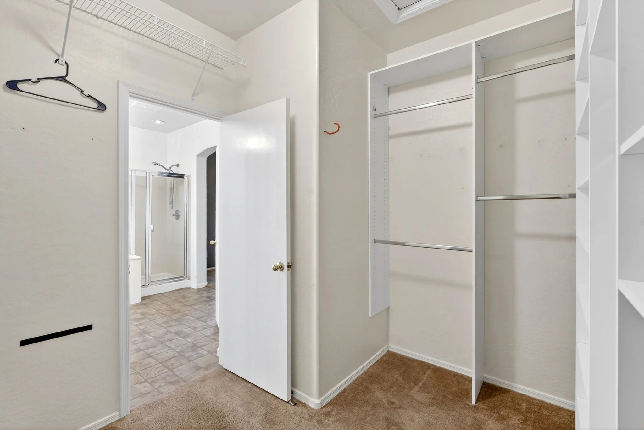 An empty walk-in closet with white shelving and rods for clothes, beige carpet, and an open doorway leading to a bathroom with a shower.