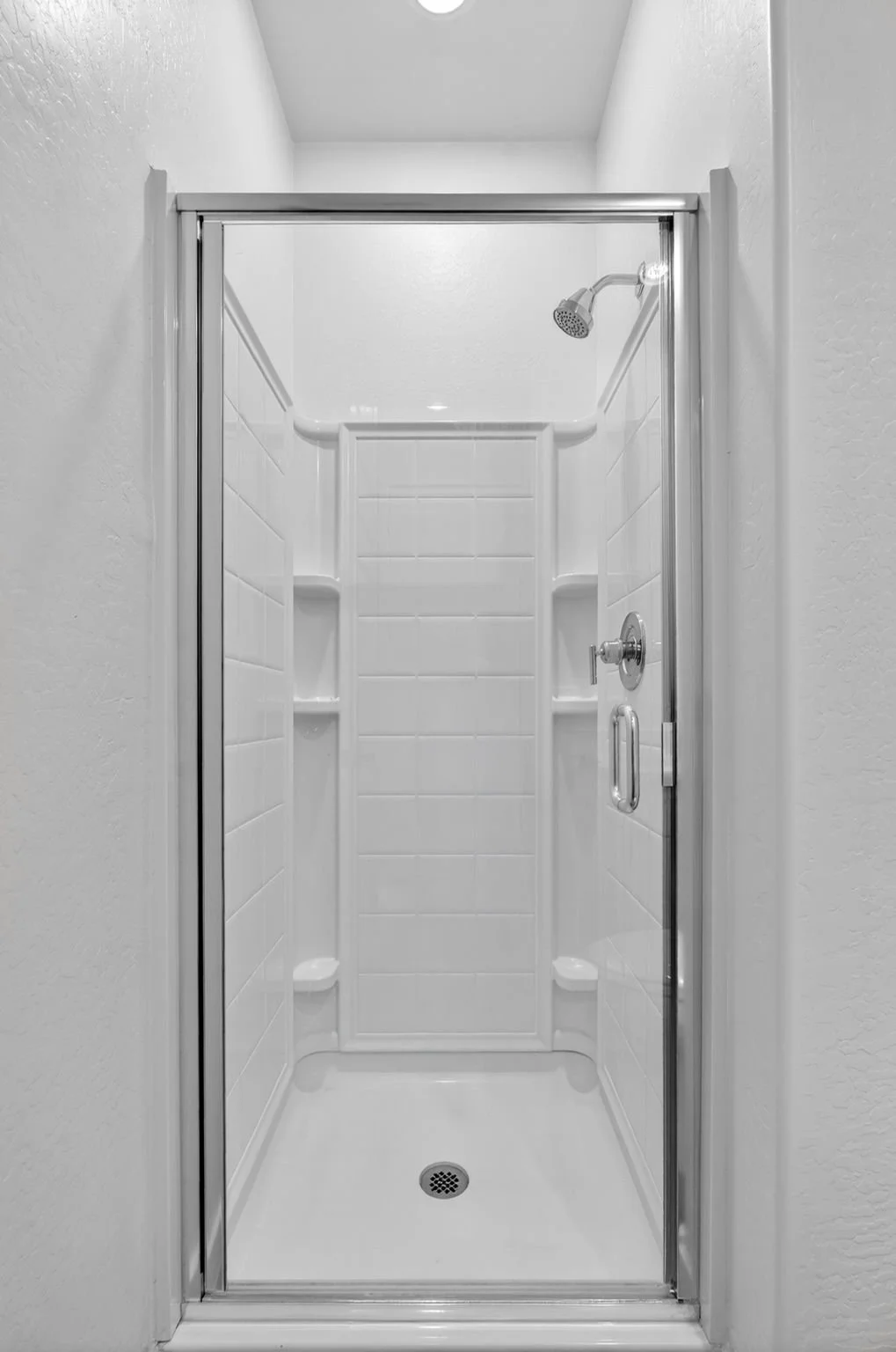 Empty white shower with built-in shelves, a handheld showerhead, and a glass sliding door.