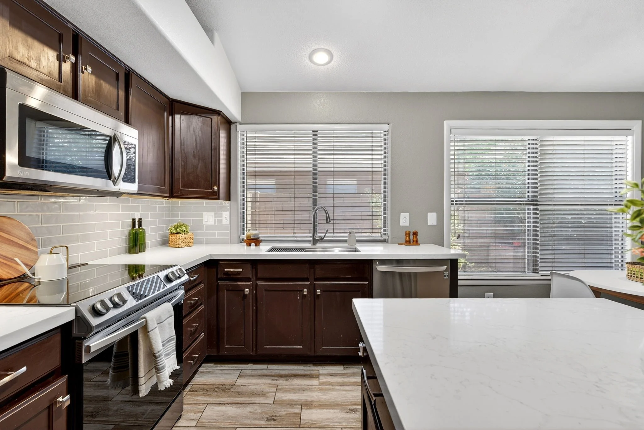 Kitchen with dark wood cabinets, white countertops, light wood plank flooring, stainless steel appliances, and two large windows with blinds providing natural light.