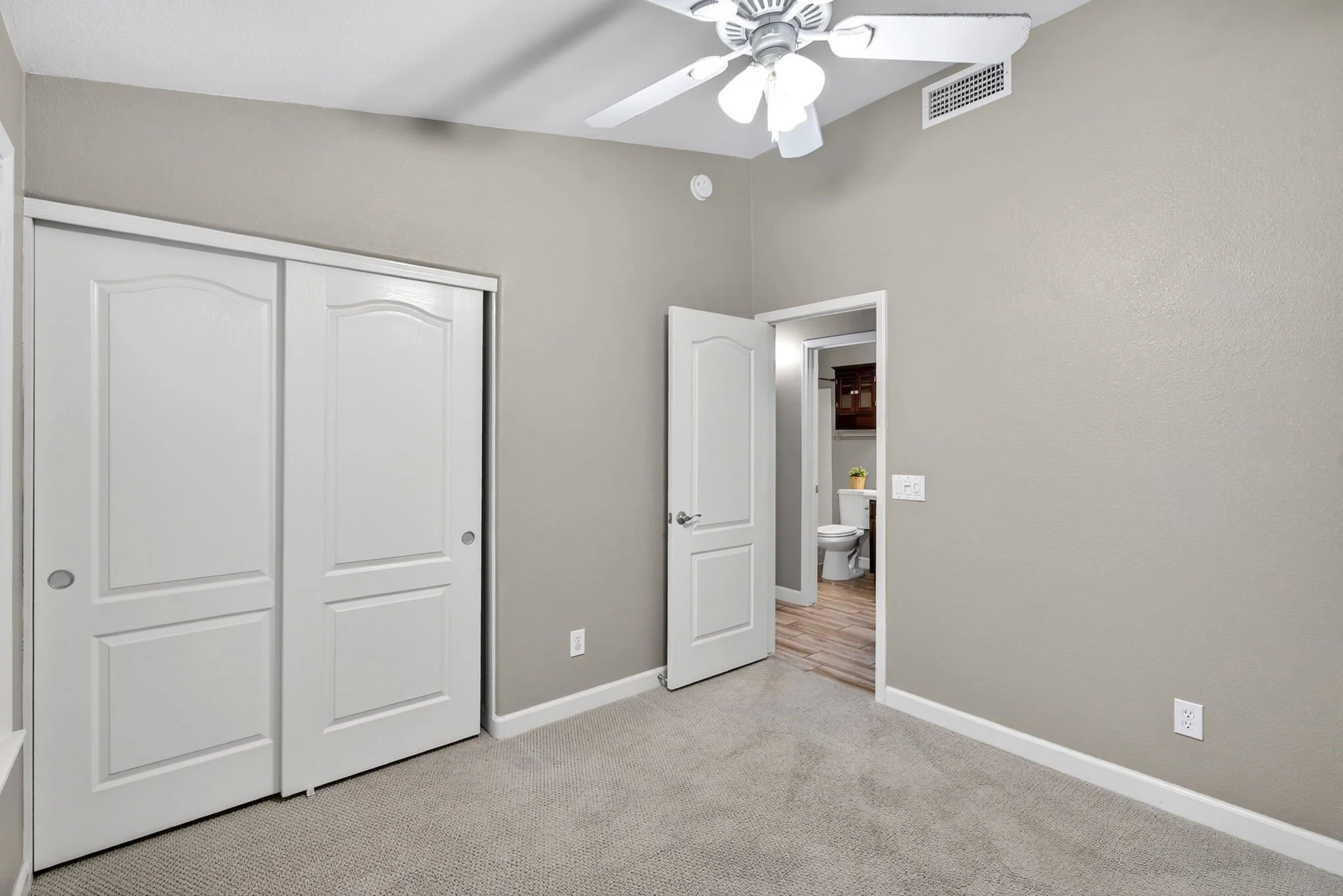 A room with neutral-colored walls, a closet with white sliding doors, and a ceiling fan with light fixtures. There is an open door leading to a bathroom with a toilet and wooden cabinets visible.