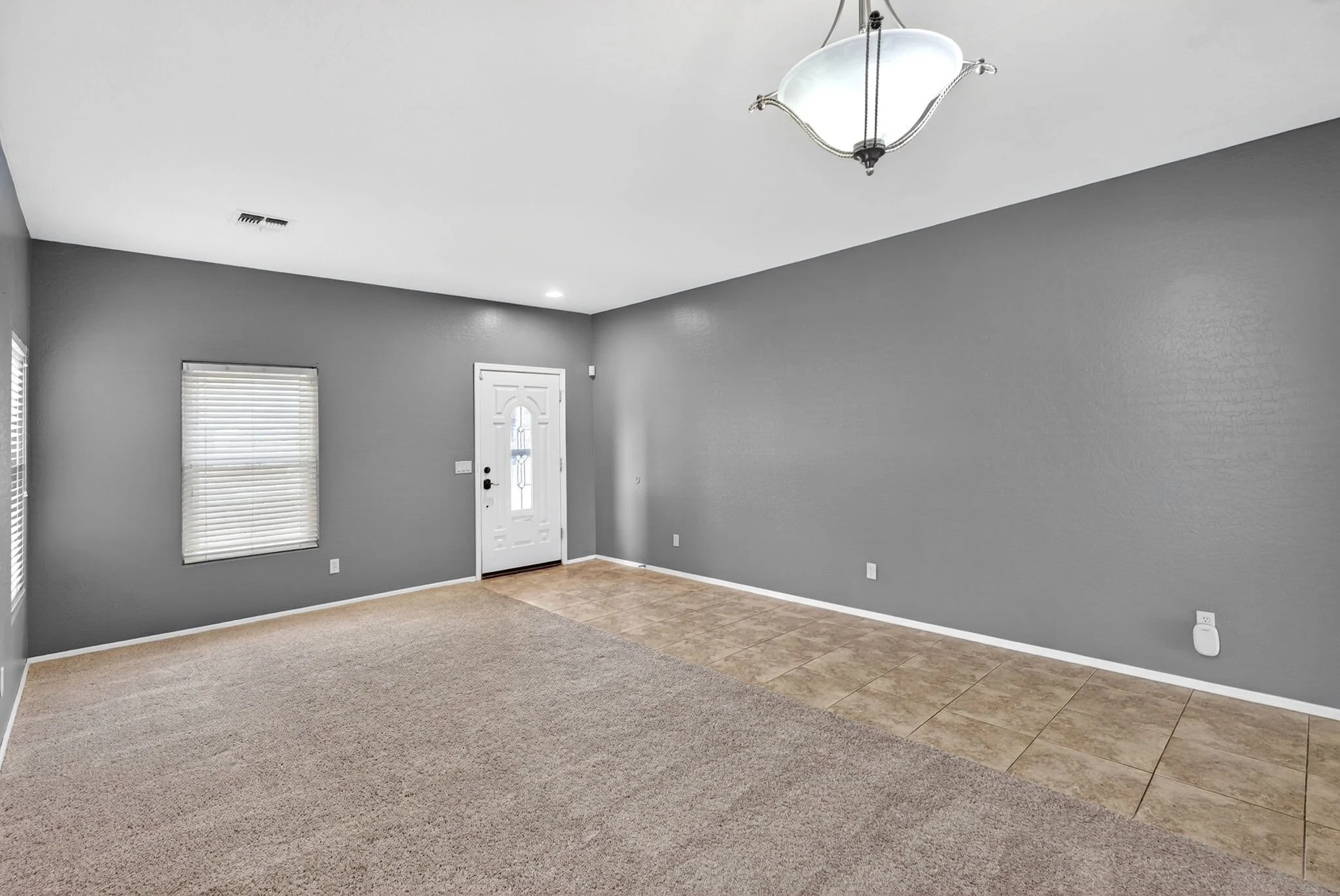 An empty living room with gray walls, a white ceiling, a door with decorative glass, a window with blinds, a beige carpeted area, and a tiled section near the wall.