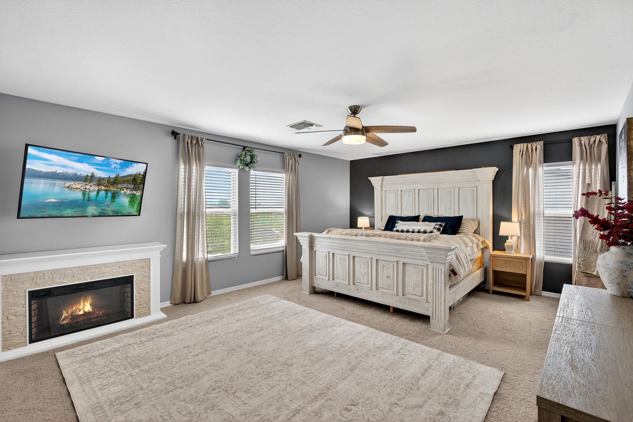 Living room with beige carpet, a white stone fireplace with a fire, a wall-mounted TV showing a lake with snow-capped mountains, a white bed frame, dark blue and patterned pillows, beige curtains, a ceiling fan, and a side table with a lamp.