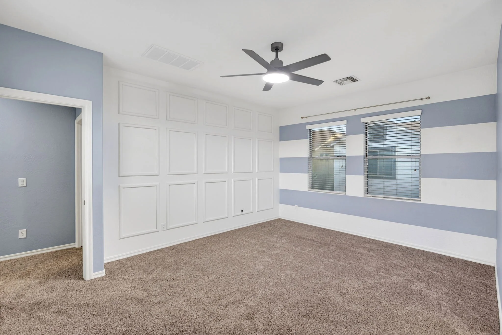 Empty room with white and light blue walls, carpeted floor, multiple windows with blinds, ceiling fan with light, and decorative white wall paneling.