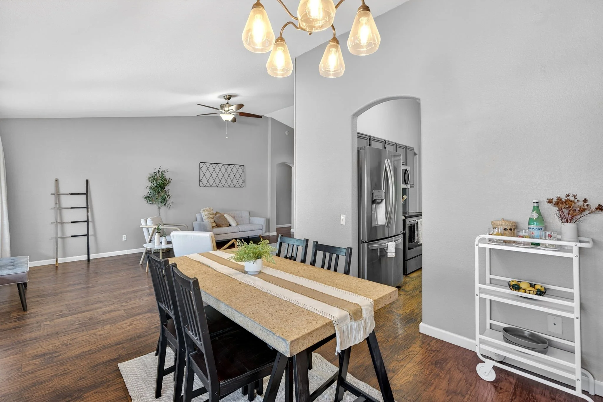 Open-concept living and dining area with a wooden dining table, black chairs, and a kitchen in the background with stainless steel refrigerator and gray cabinets. There is a white cart with kitchen items and a table with a central potted plant. The r