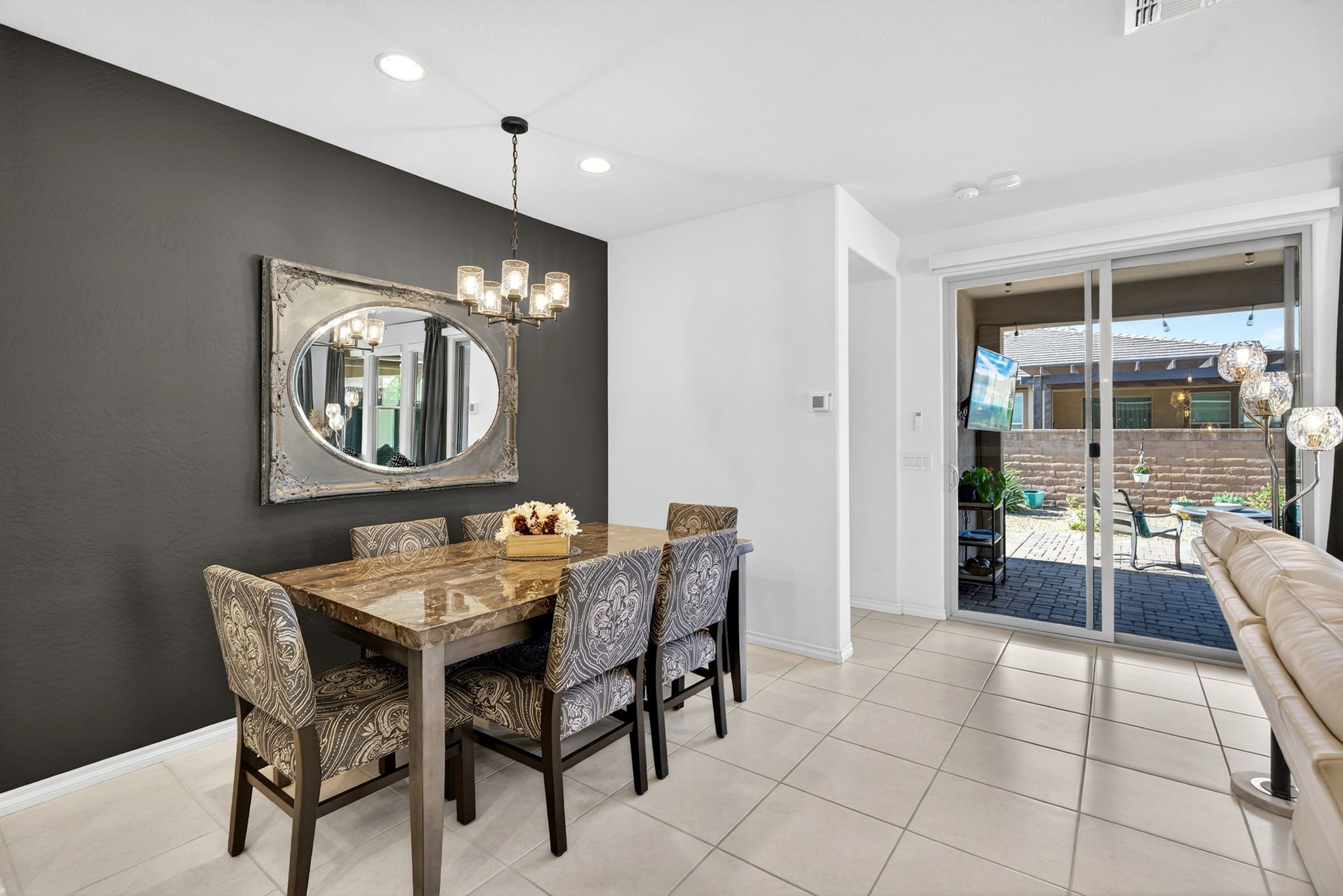 Dining area with a marble-top table, patterned chairs, and a large mirror on a dark painted wall, next to sliding glass doors leading to an outdoor patio.