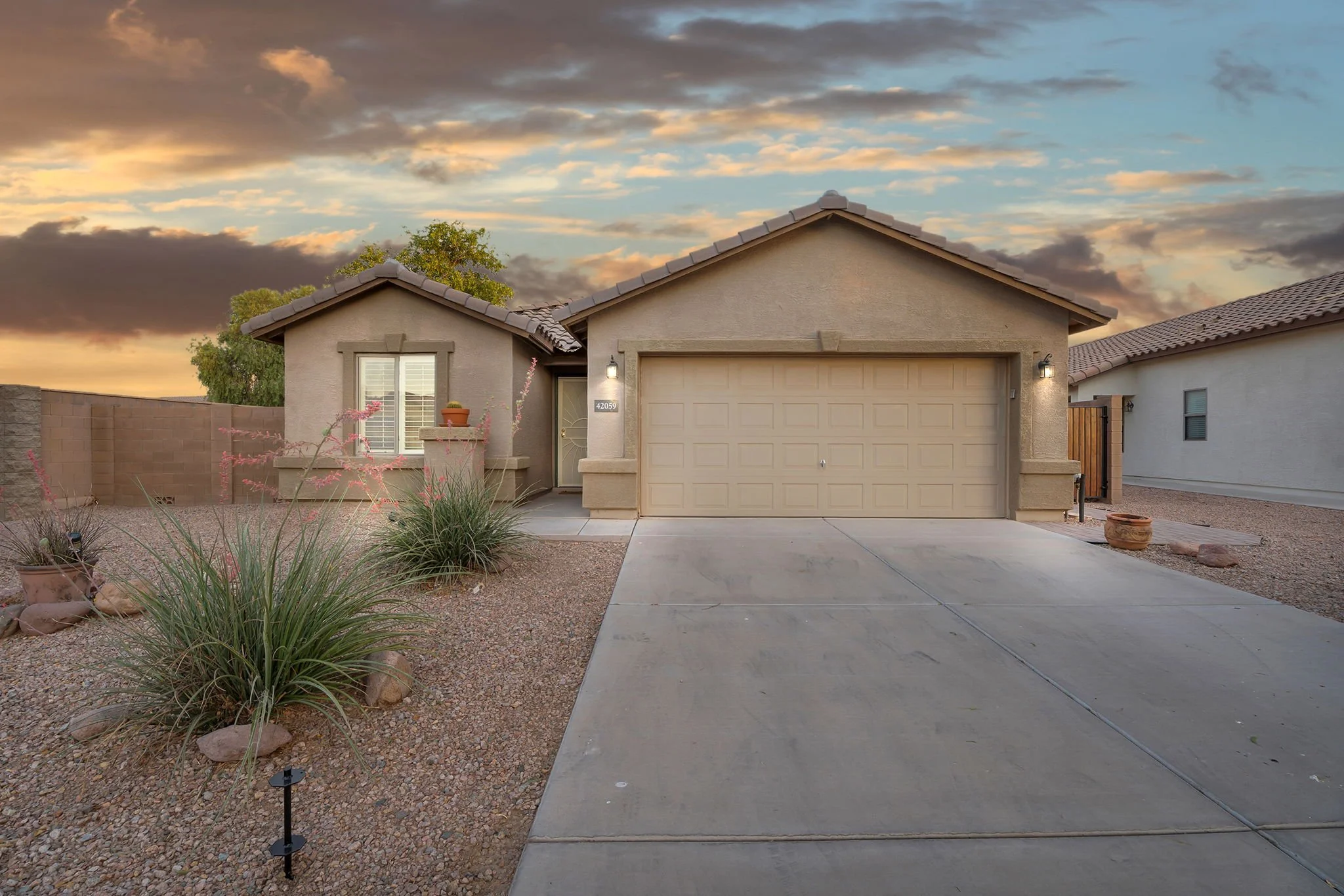 A single-story beige house with a closed garage door, front window with shutters, and front door with a glass panel, set against a cloudy sunset sky, with desert landscaping including rocks and drought-tolerant plants in the yard.
