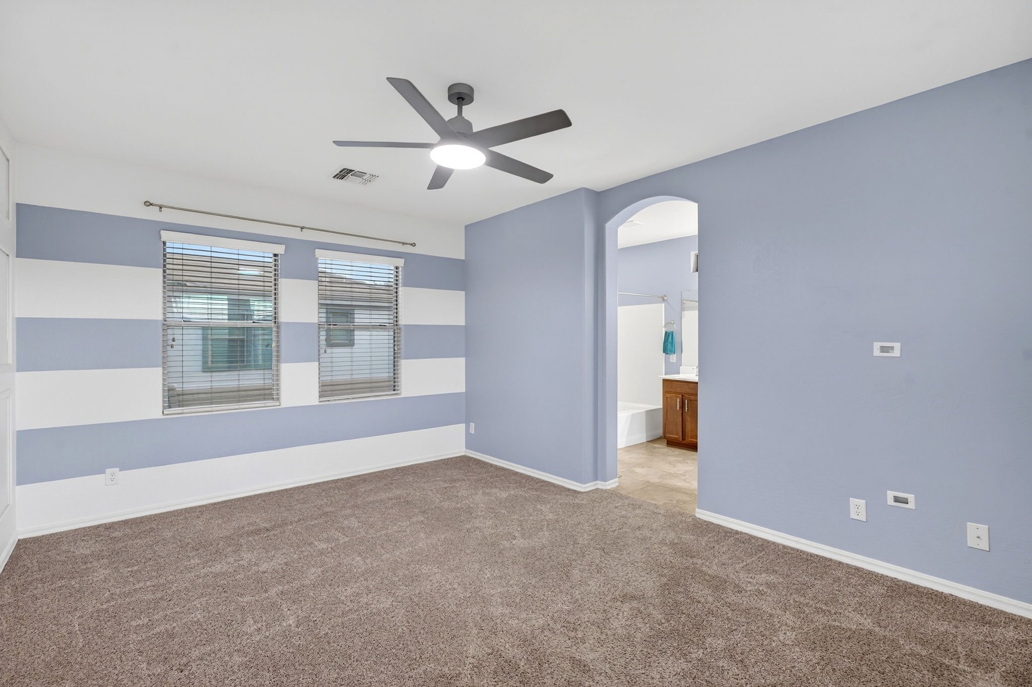 An empty room with striped blue and white walls, two windows with blinds, a ceiling fan with light, carpeted floor, and a doorway leading to a bathroom with wooden cabinets.