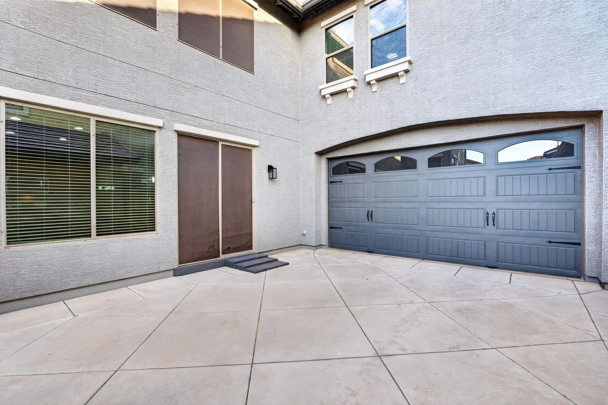 A view of a modern house's backyard with a gray driveway, a garage door, a sliding door with blinds, and two windows with decorative white corbels beneath them.