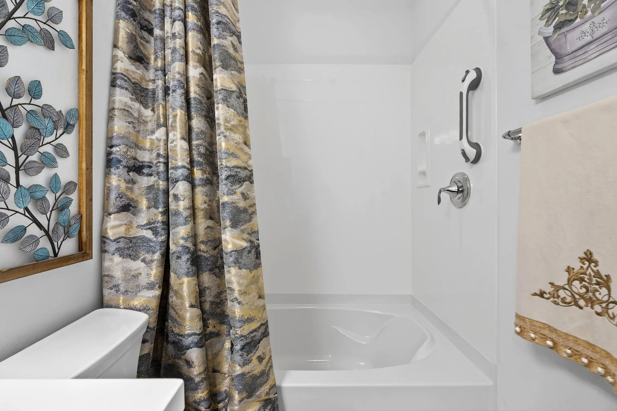 A bathroom with a white bathtub, a shower curtain with an abstract pattern in shades of gray, gold, and blue, a decorative wall art piece with metallic leaves, a white toilet, a white vanity with a marble top, and a framed floral painting on the wall