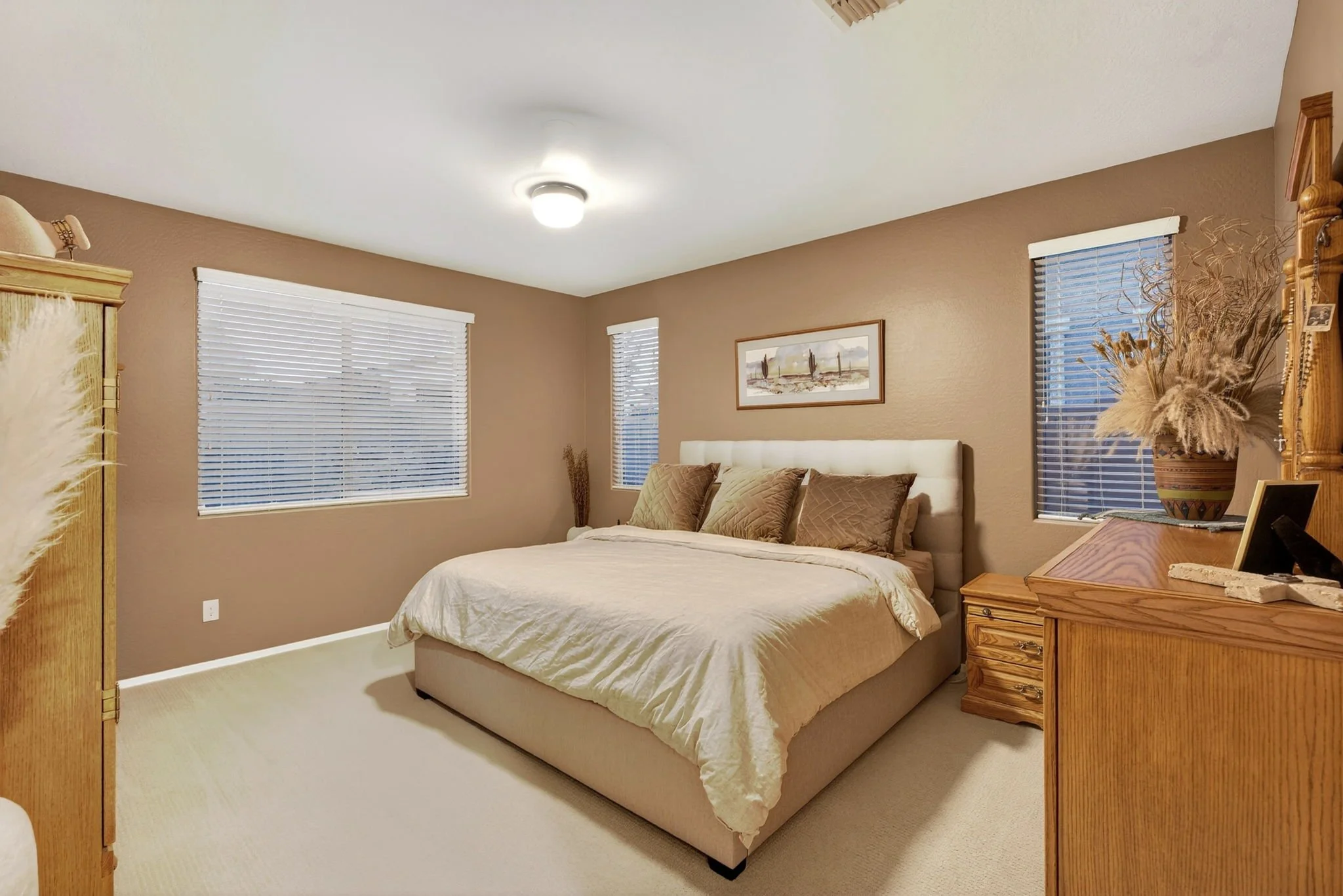 A cozy bedroom with a large bed, brown and beige color scheme, two windows with blinds, a picture of cacti on the wall, and wooden furniture including a dresser and nightstand with a decorative vase and picture frame.