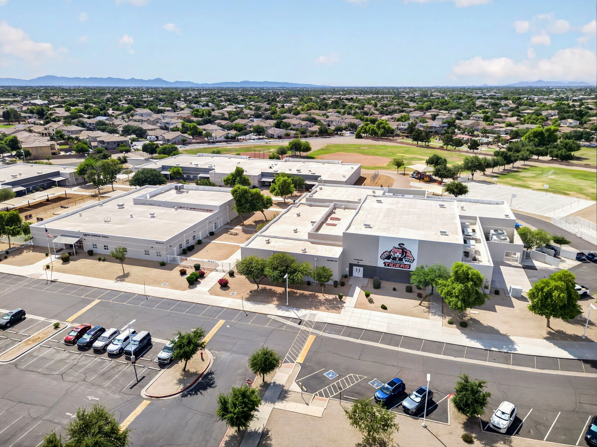 Aerial view of a schoolyard with white buildings, parking lot, football field, and surrounding residential neighborhood with trees and mountains in the distance.