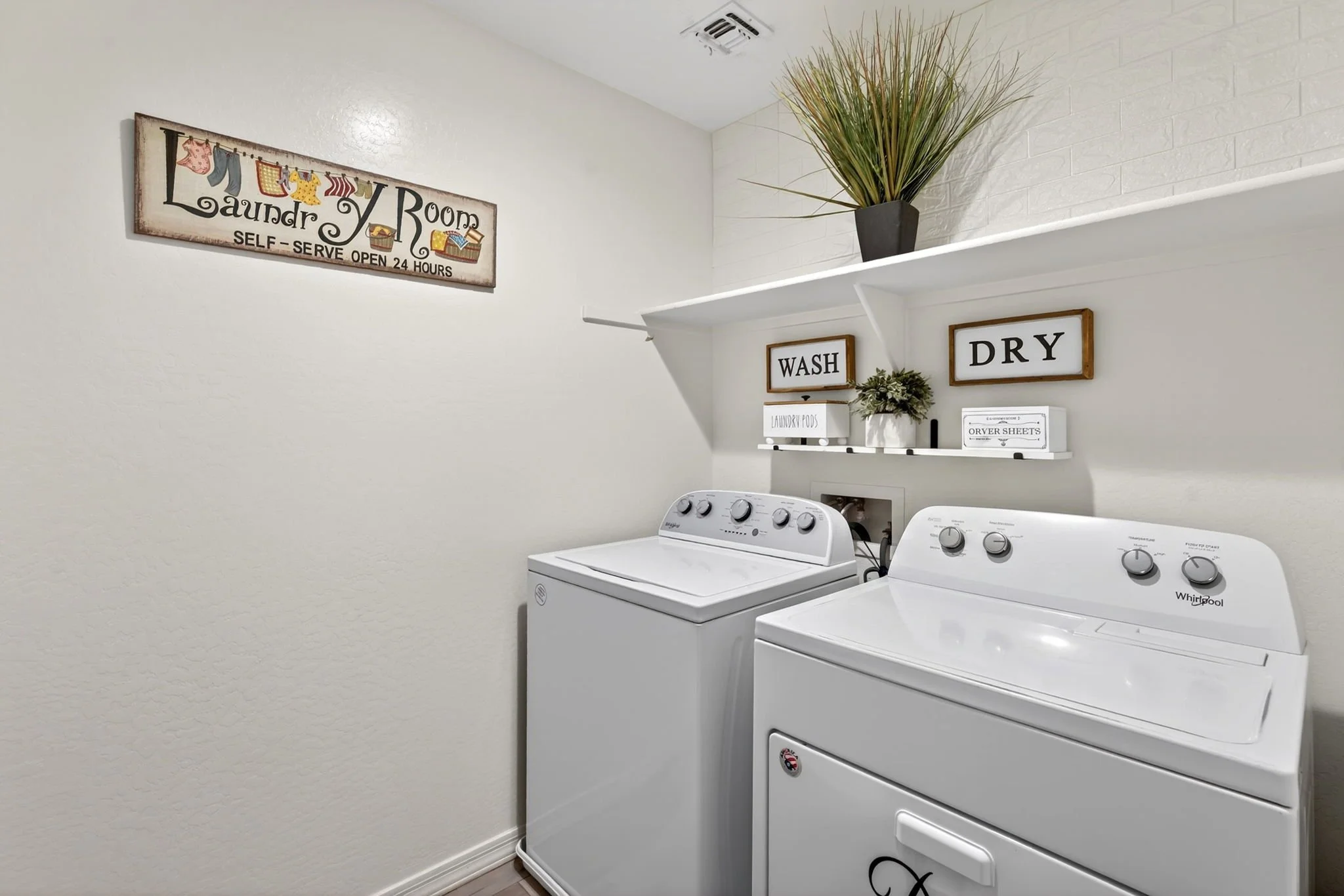 A laundry room with a white washing machine and dryer, a shelf with a potted plant and laundry supplies, and decorative signs that say 'Wash' and 'Dry' on the wall.
