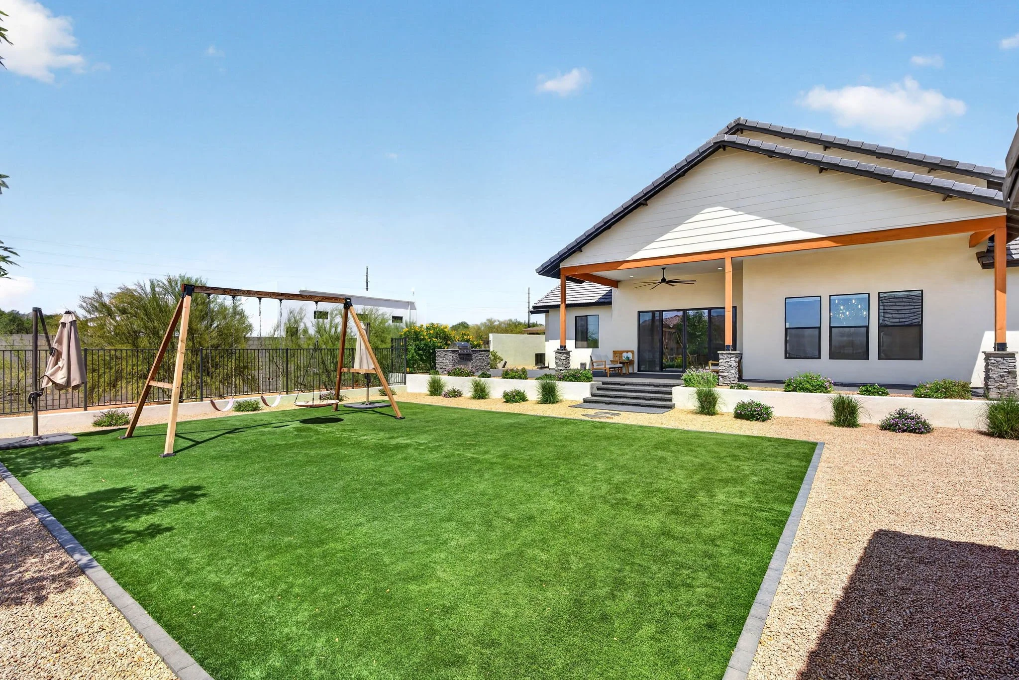 Backyard with swingset, lawn, patio, house with roof and large windows, and landscaped plants.