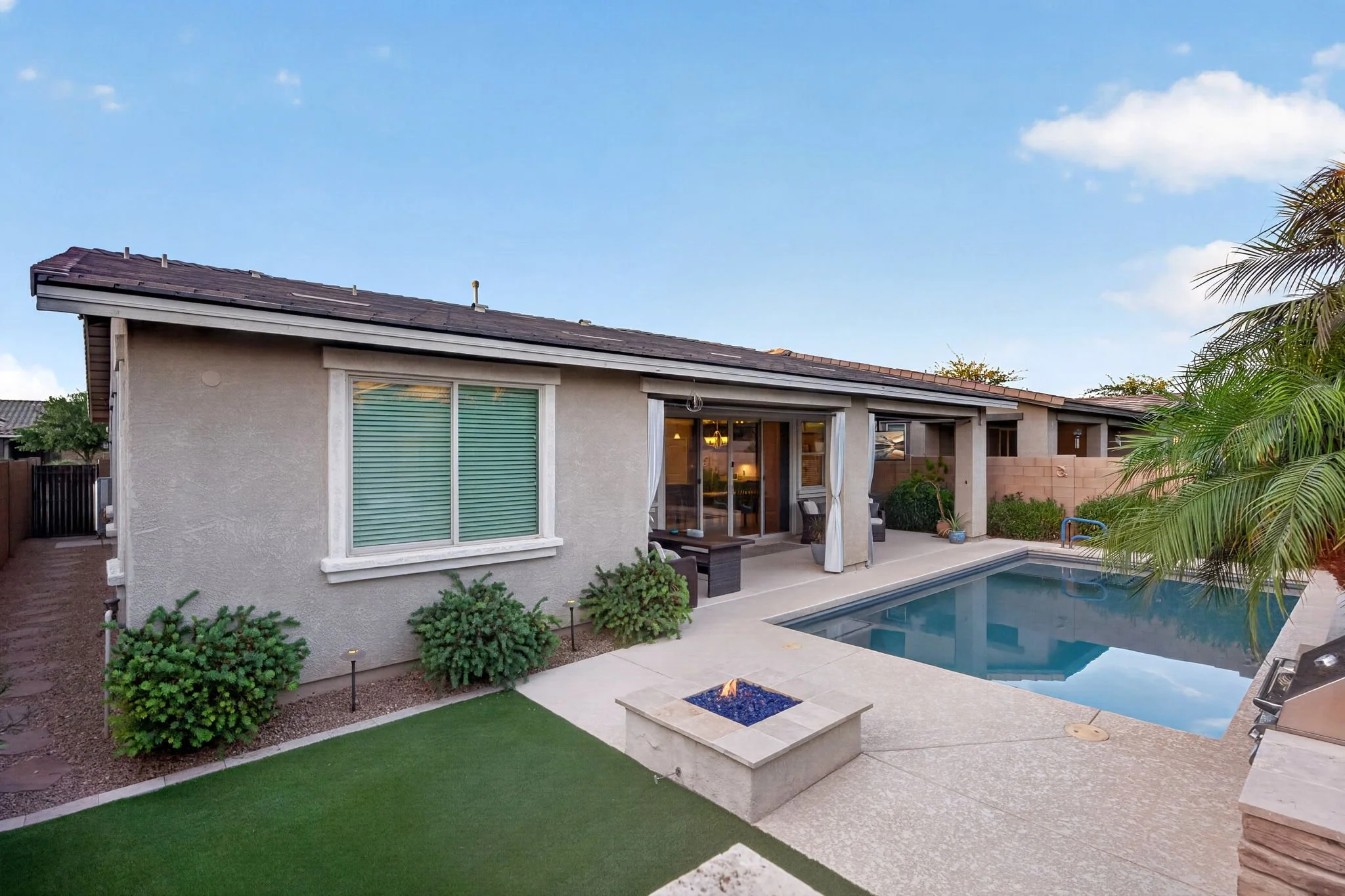 Backyard view of a house with a swimming pool, patio area, green lawn, and landscaping plants.