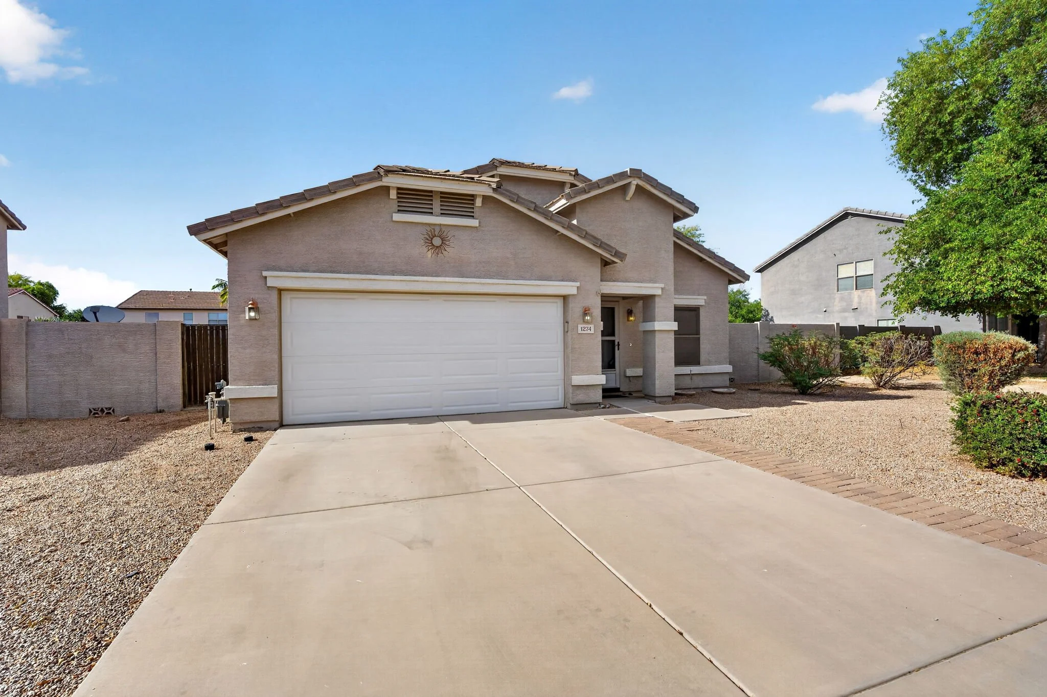 A two-story house with a white garage door, light-colored exterior walls, and a small front yard with bushes. There is a sidewalk and a gravel area in the front yard. A tree with green leaves is on the right side of the house. The sky is blue with a 