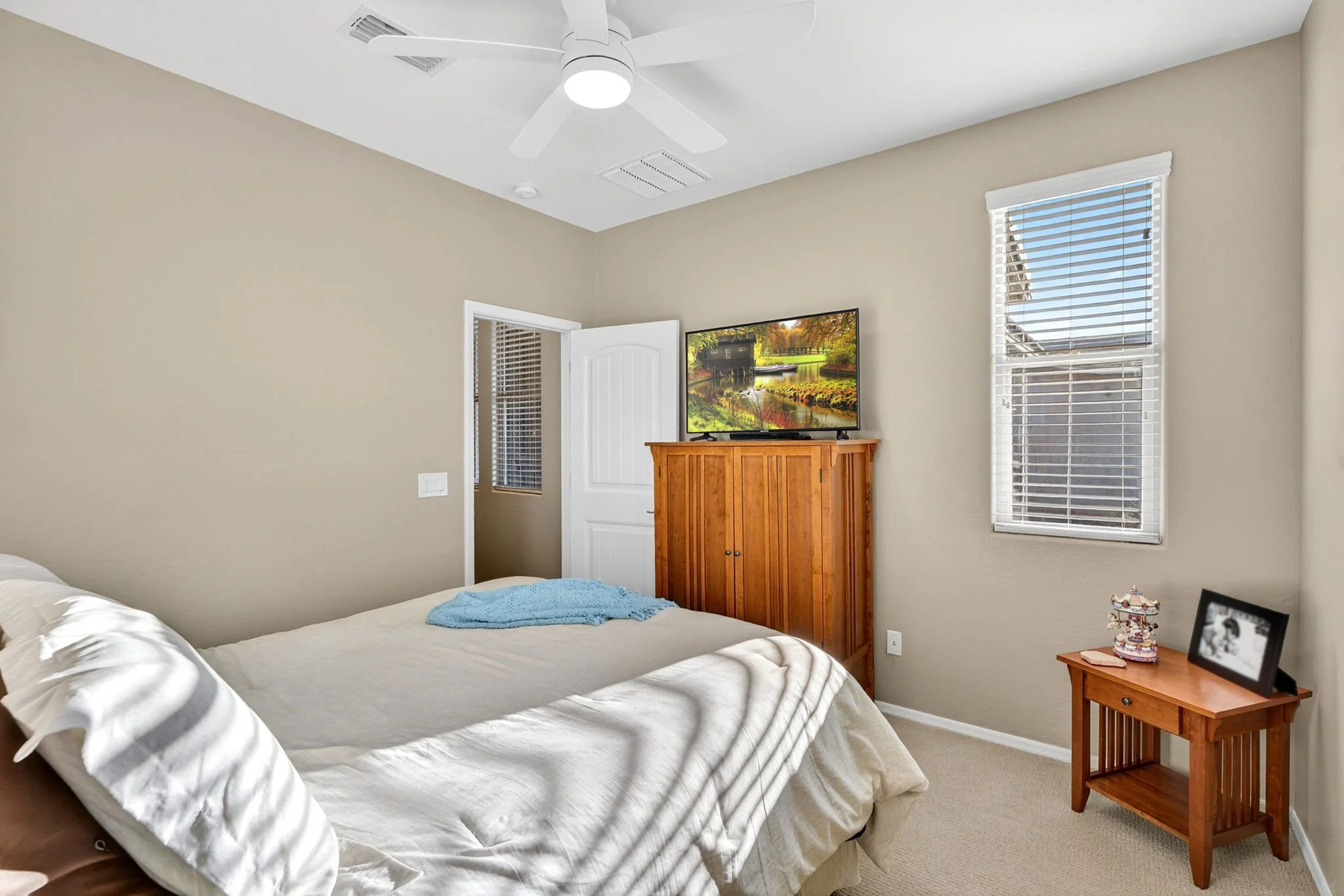 A bedroom with a bed in the foreground, a wooden dresser with a TV on top, a window with blinds, and a nightstand with a framed photo and decorative items.