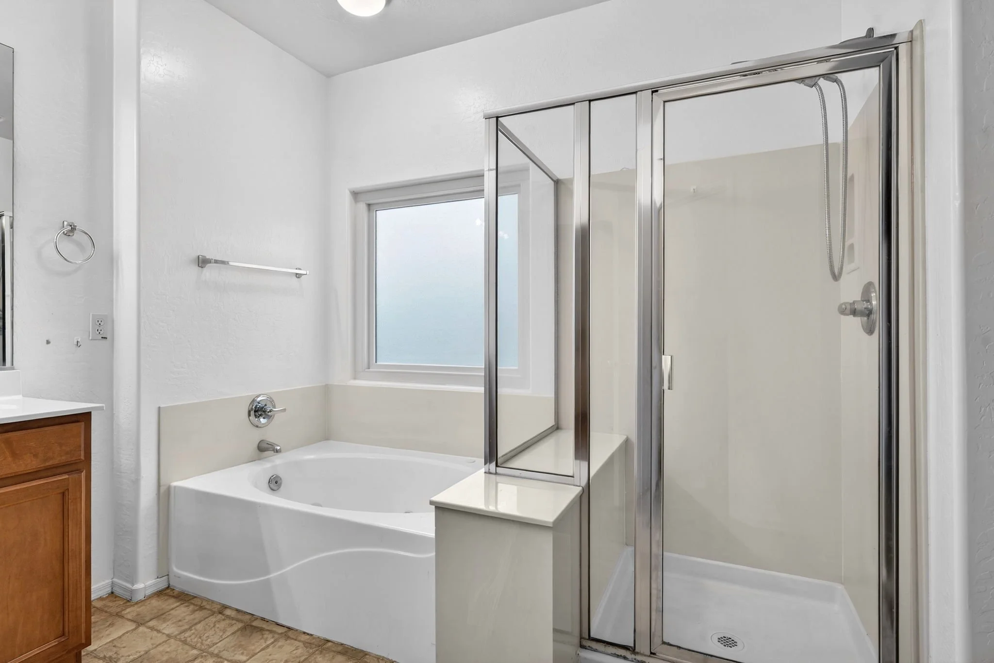 A bathroom with a bathtub, a glass shower stall, and a window. The bathtub is built into the corner, with a silver faucet and handles. The glass shower stall has a sliding door, with a metal showerhead. There are white walls, a towel bar, and an elec