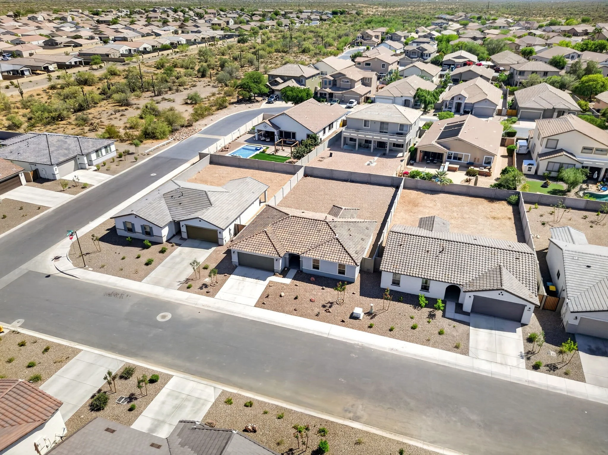 An aerial view of a suburban neighborhood with houses, lawns, trees, a pool, and desert landscape.