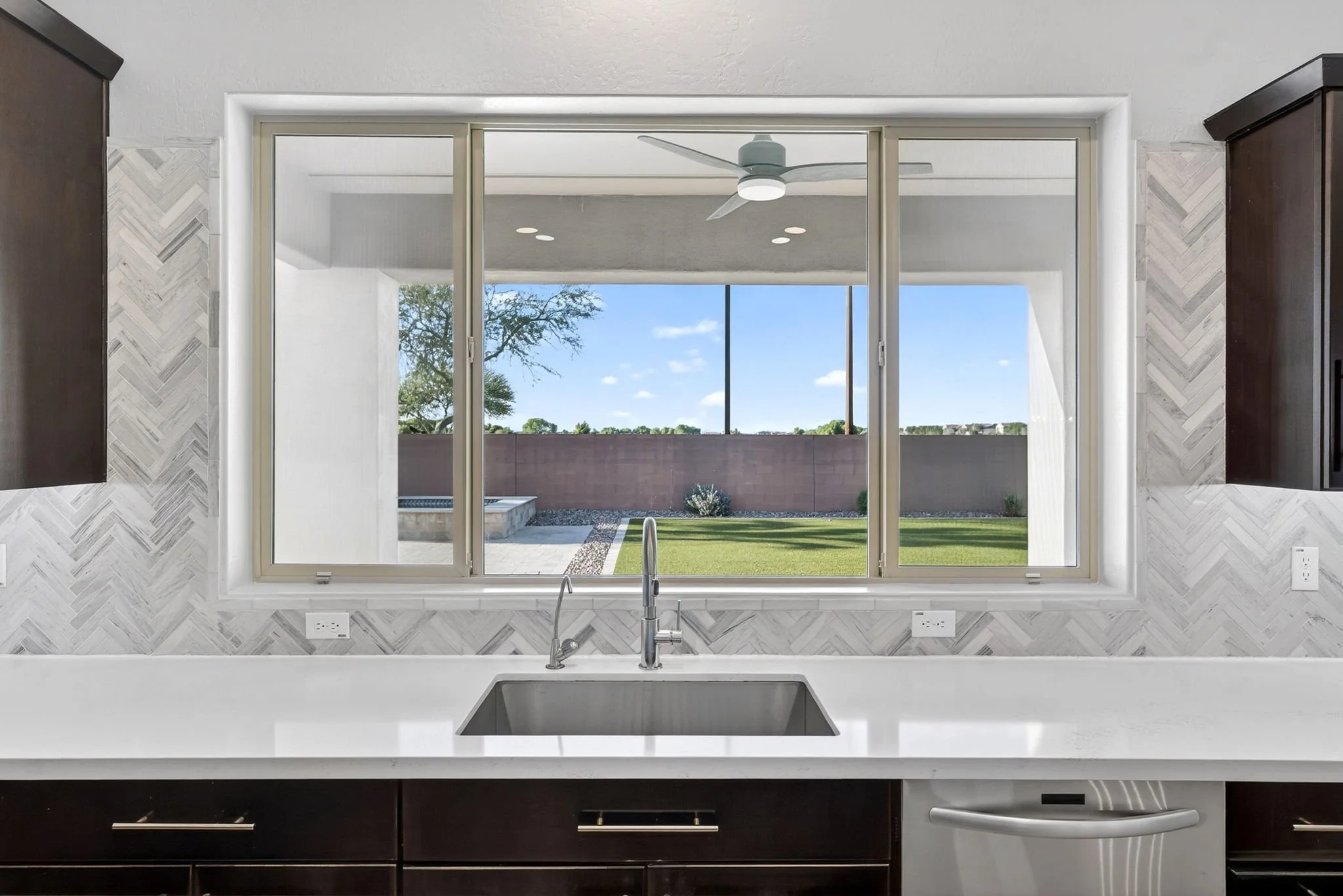 Modern kitchen with large window above sink showing outdoor patio, green lawn, and blue sky, with dark cabinets below and patterned wall tiles.