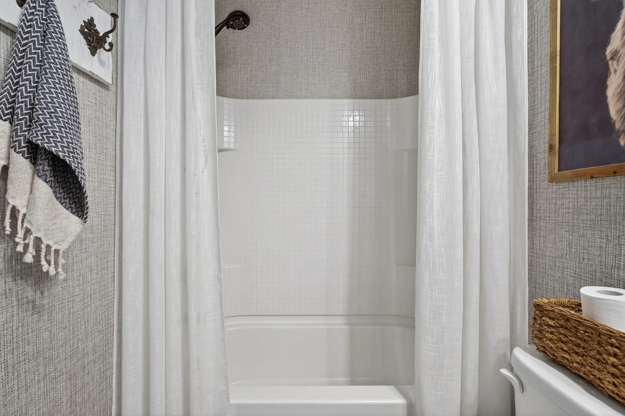 Bathroom with a bathtub enclosed by sheer white curtains, gray textured wall, and various decorative items including towels, a framed chalkboard, and a wicker basket.