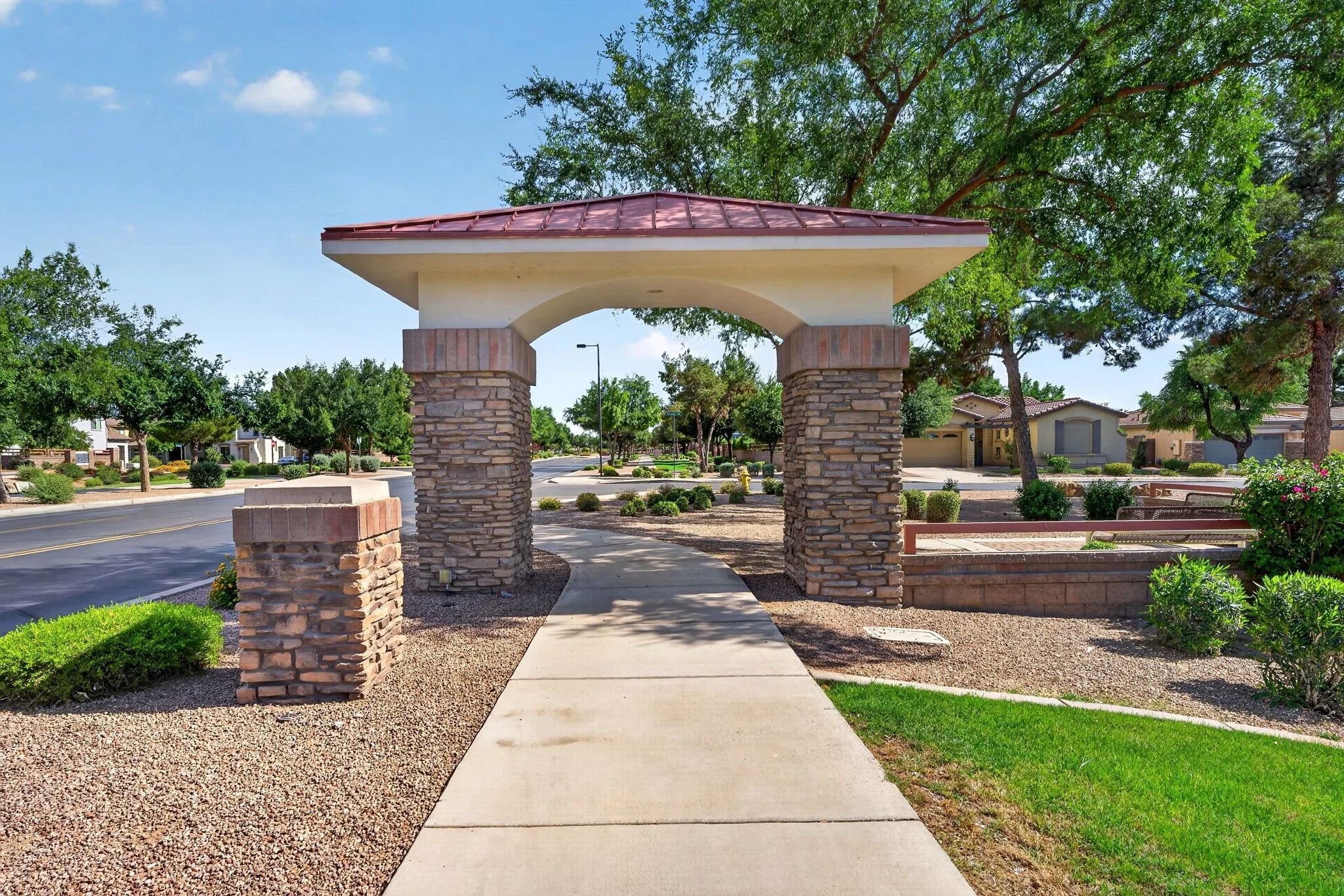 A sidewalk leading through an archway structure with stone pillars, located in a suburban neighborhood with green trees and houses in the background under a clear blue sky.