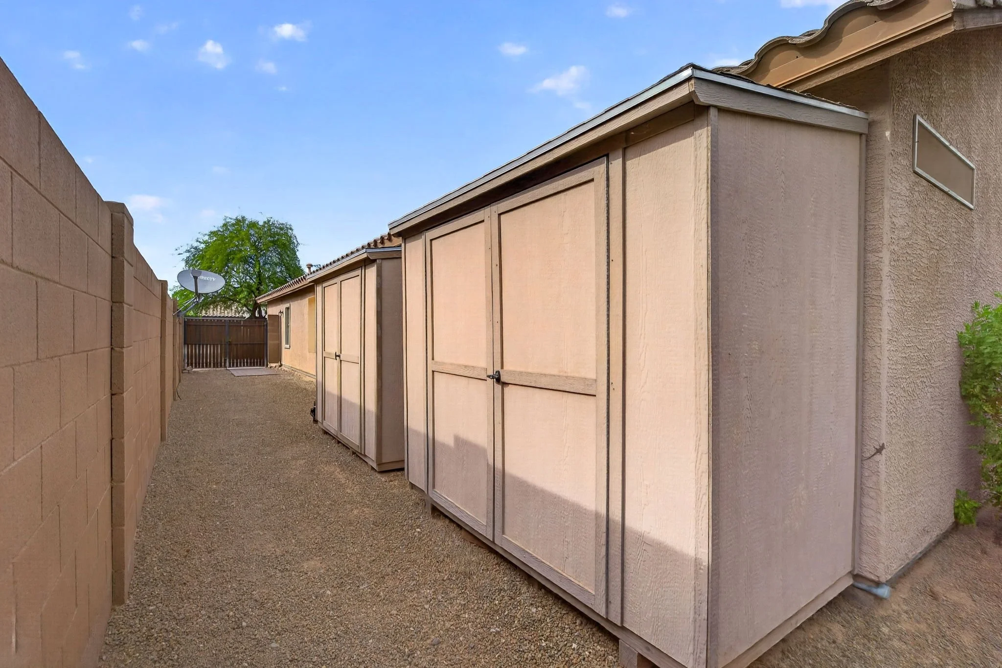 A narrow backyard alley between a brick wall on the left and a beige stucco house on the right, with a gravel ground, a satellite dish mounted on the brick wall, a tree in the background, and a gate at the end of the alley.
