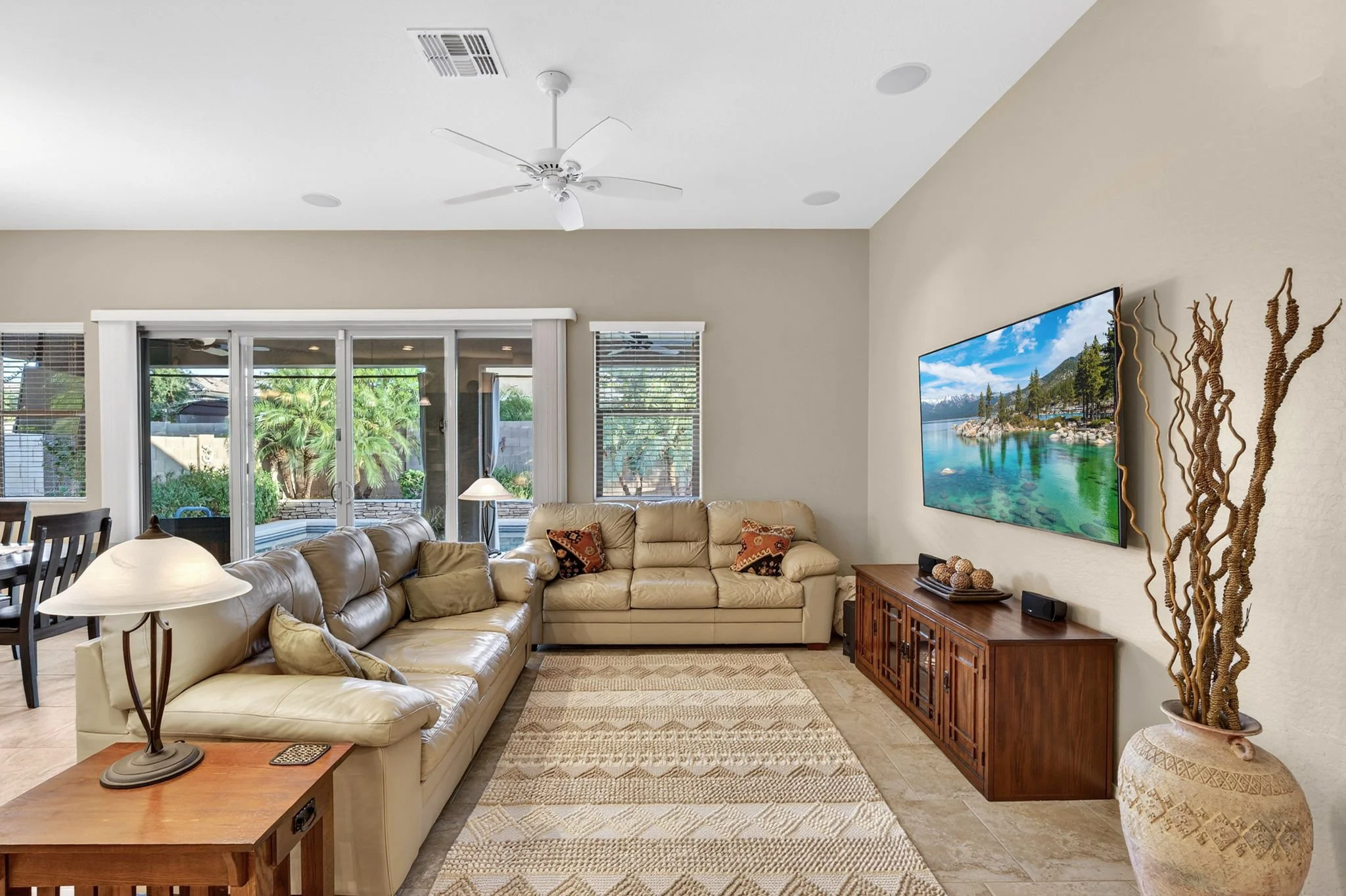 Living room with beige sectional sofa, wooden side table, lamp, decorative pillows, large window, sliding glass door leading outside, wall-mounted flat screen TV displaying mountain and lake scenery, and tall decorative twisted wood sculpture.
